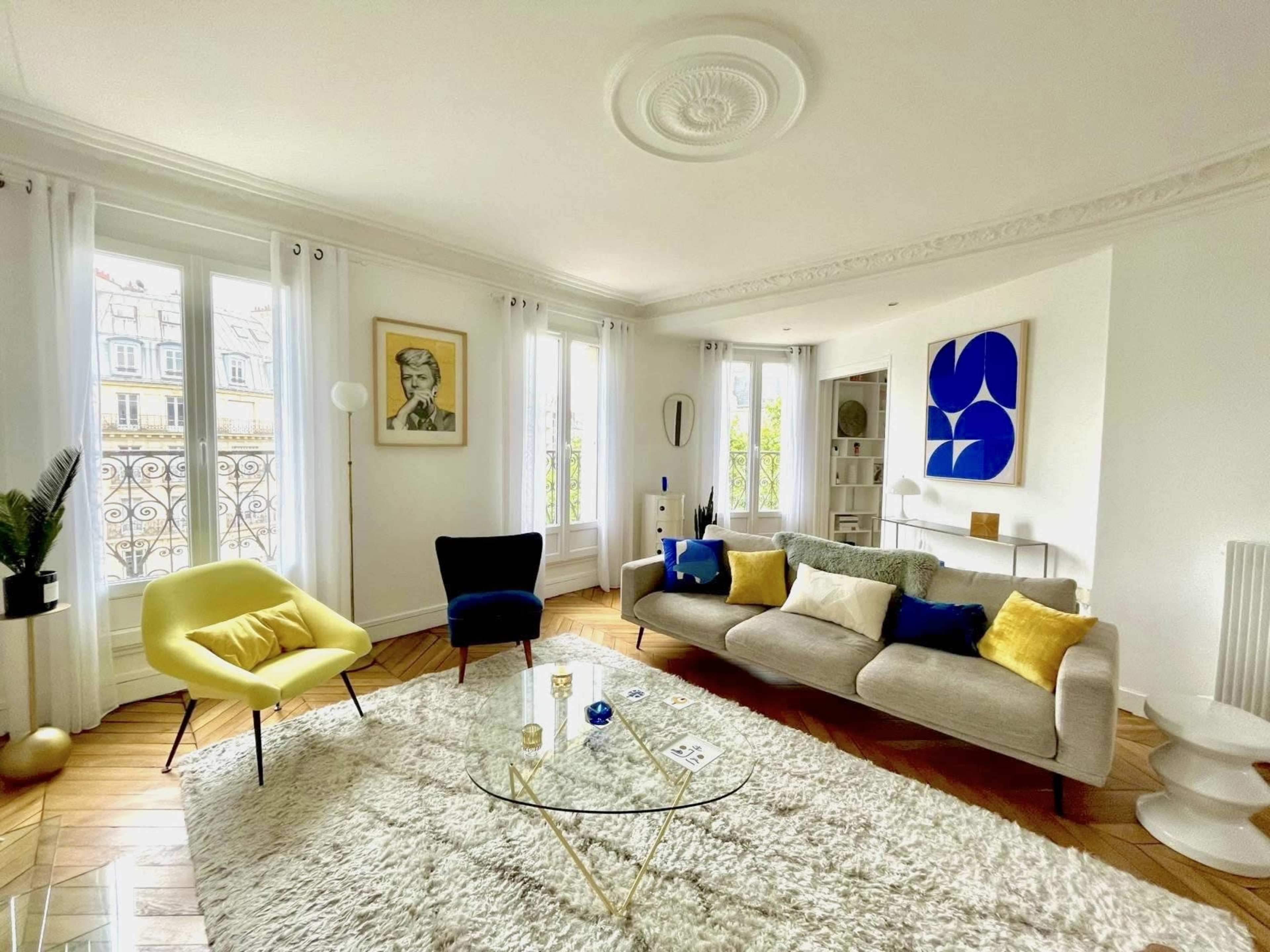 A bright, modern living room featuring a glass coffee table, a plush rug, and colorful seating arrangements near large windows.