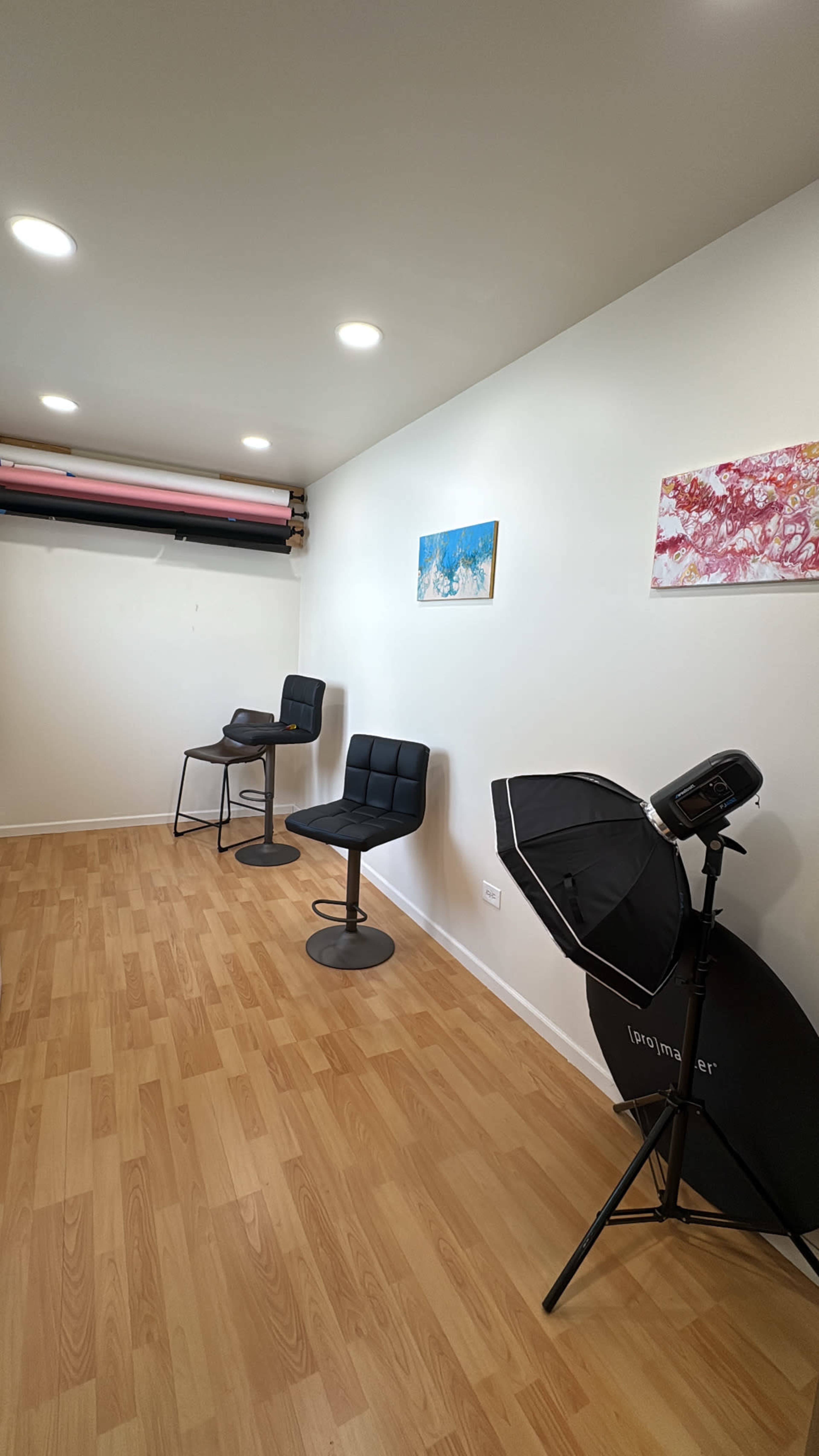 The image shows a minimalist studio space featuring two black chairs, a metal table, a lighting umbrella, and pastel artwork hanging on the walls.