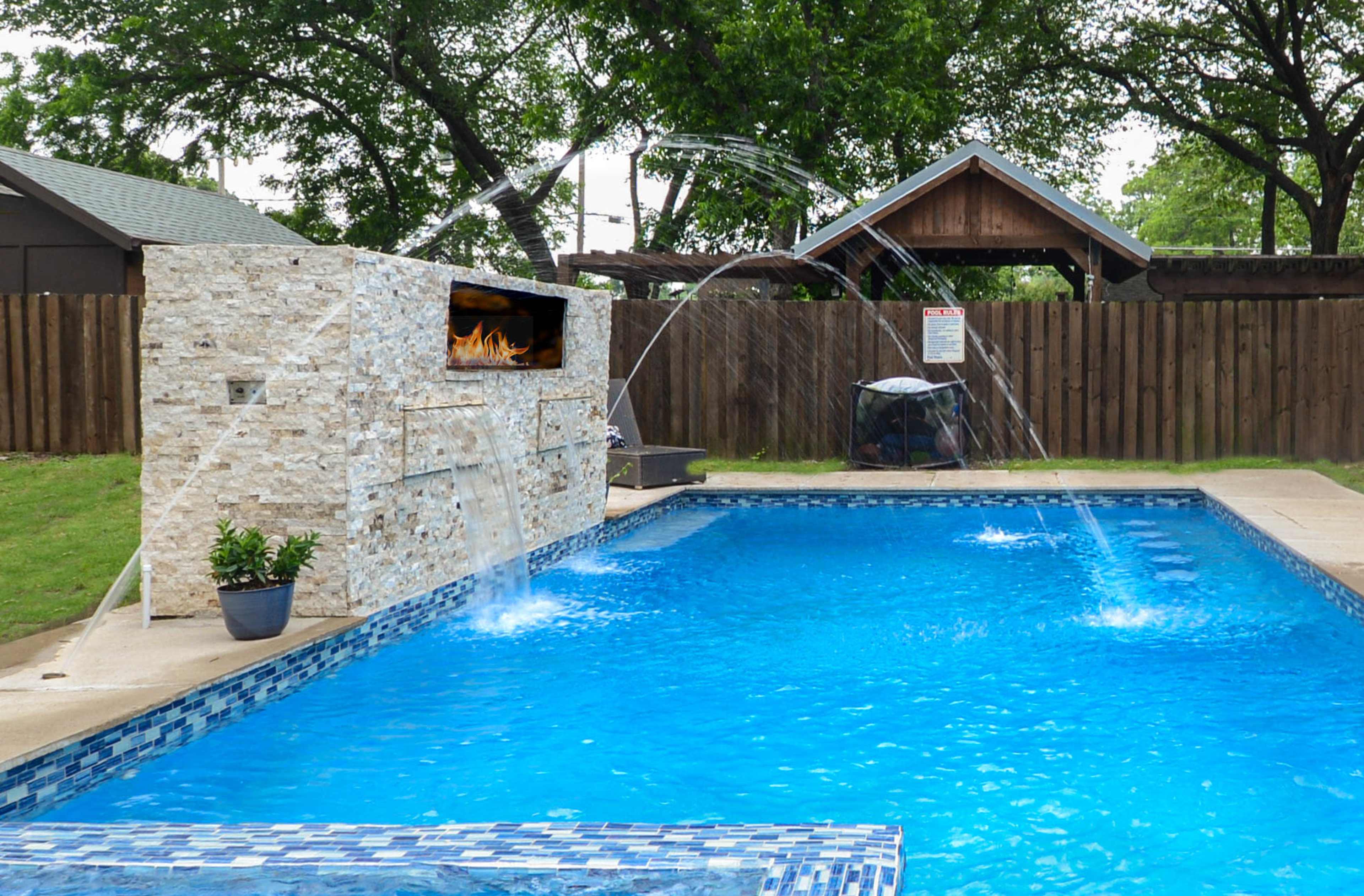 The image shows a backyard pool with a stone wall featuring a television and several water jets spraying into the water.