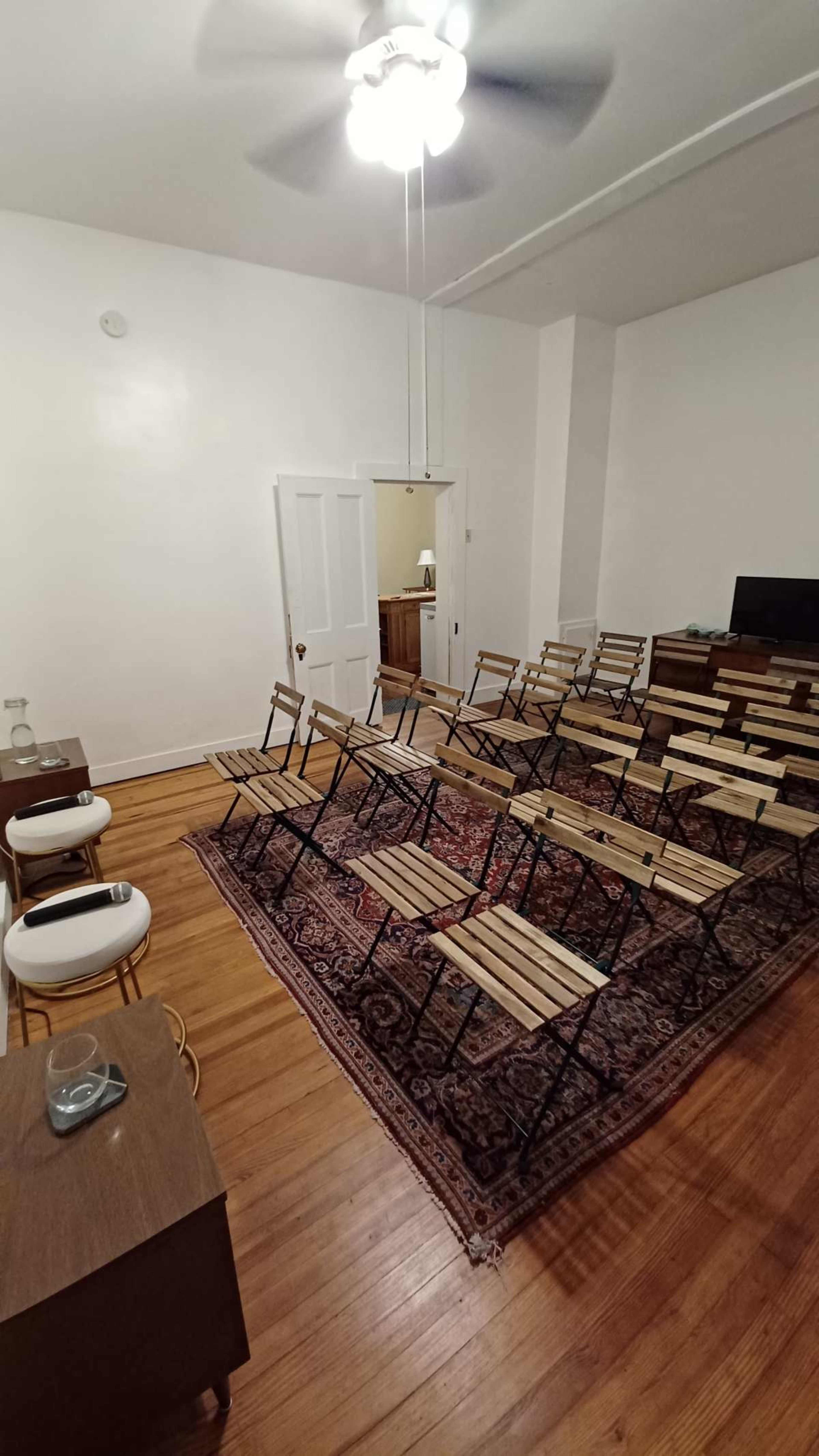 The image shows a sparsely furnished room with a wooden floor, equipped with a row of folding chairs arranged in a classroom setup, a small table, and a door leading to another area.