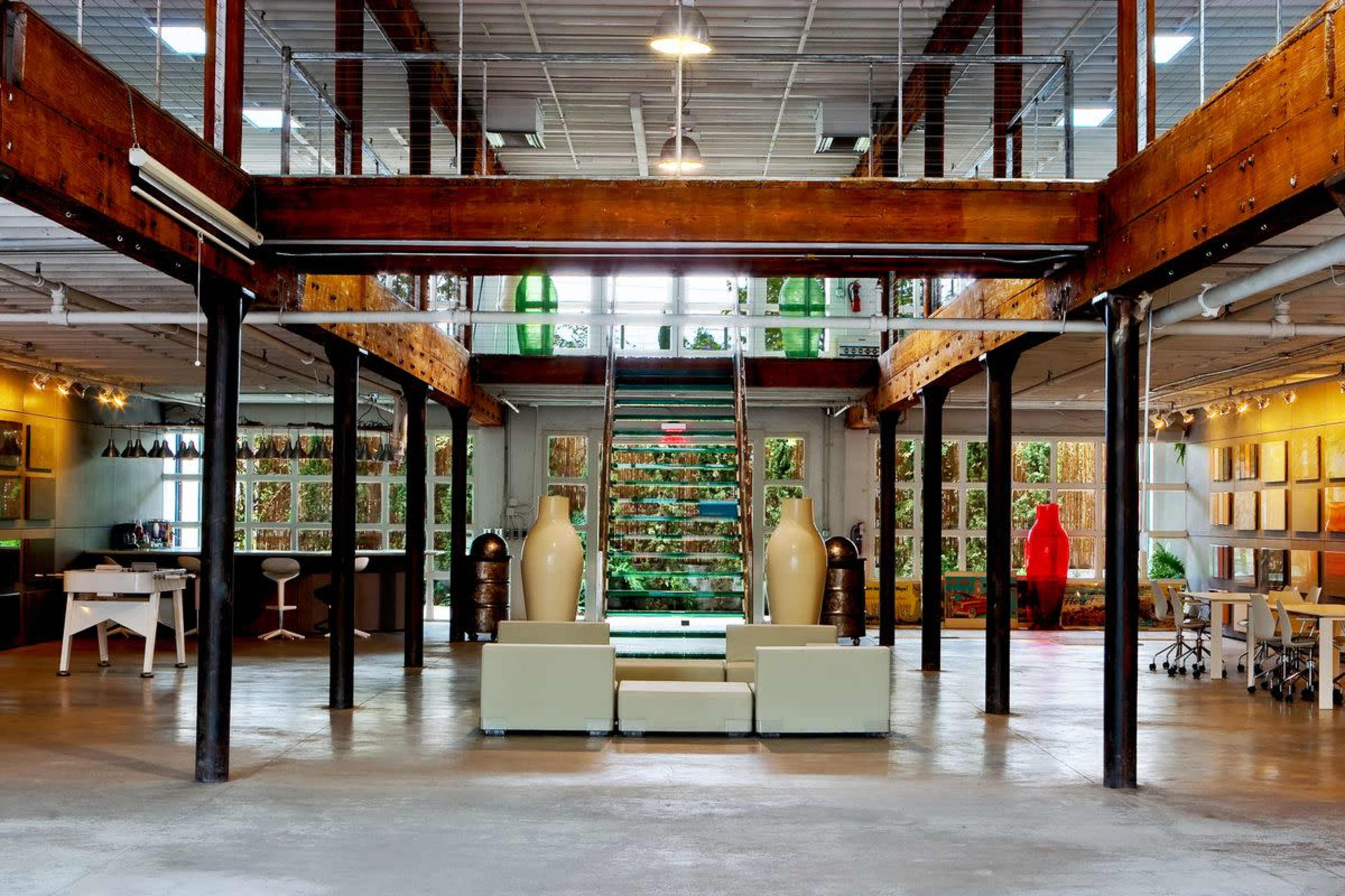 The image shows a spacious office interior with a high wooden-beamed ceiling, large windows, and a staircase leading to an upper level, featuring modern furniture and decorative vases.