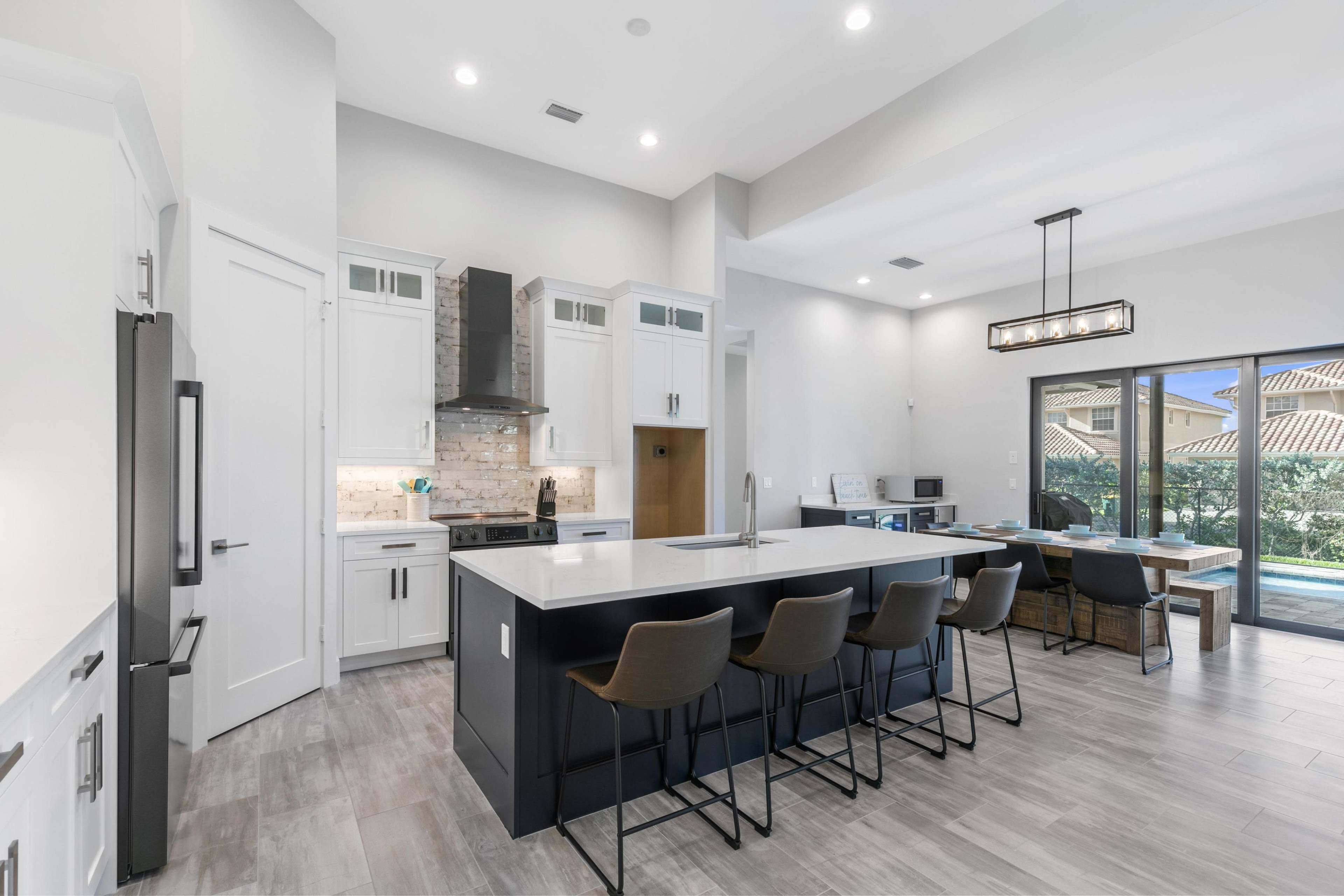 A modern kitchen with a large island, black bar stools, white cabinetry, and a view of an outdoor patio through sliding glass doors.