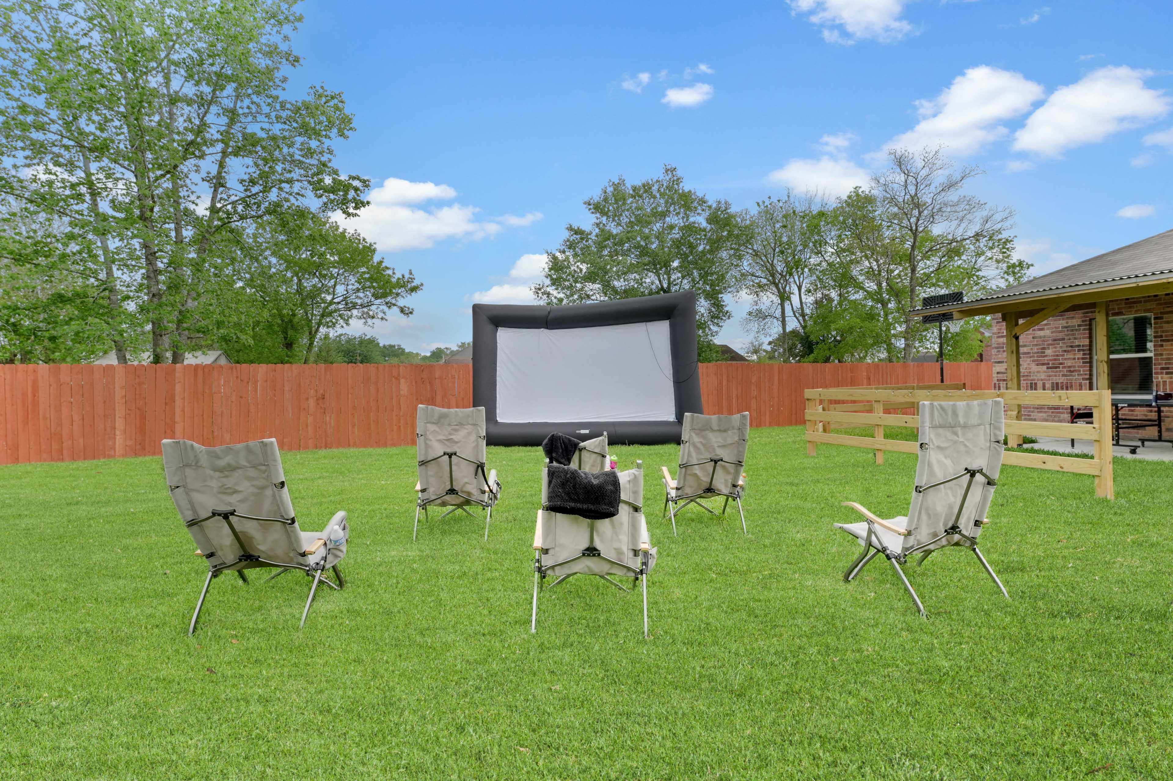 A row of six folding chairs is arranged in a circle on a grassy lawn facing an inflatable outdoor movie screen.