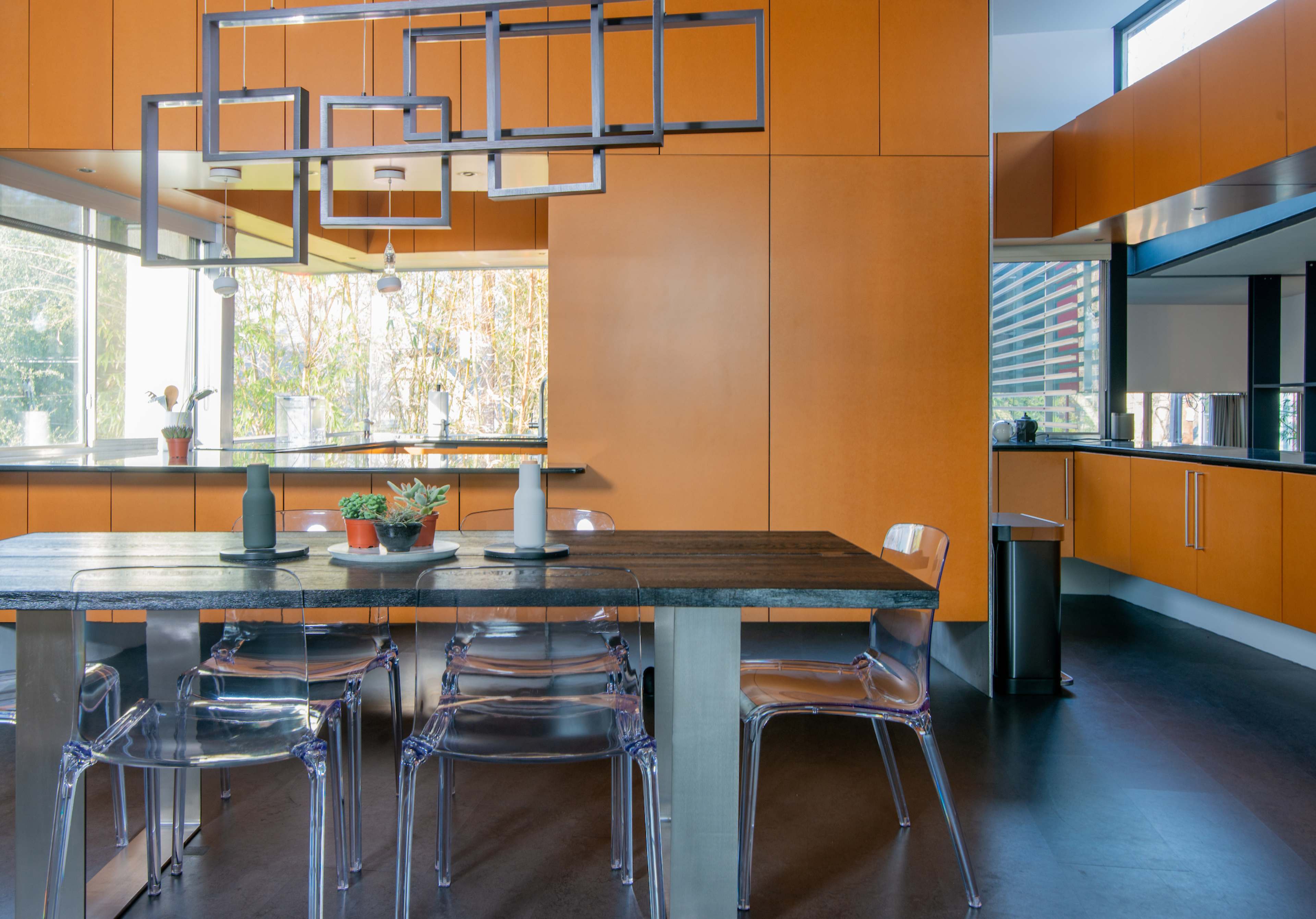 A modern kitchen features a wooden dining table surrounded by clear acrylic chairs, with orange cabinetry and large windows letting in natural light.