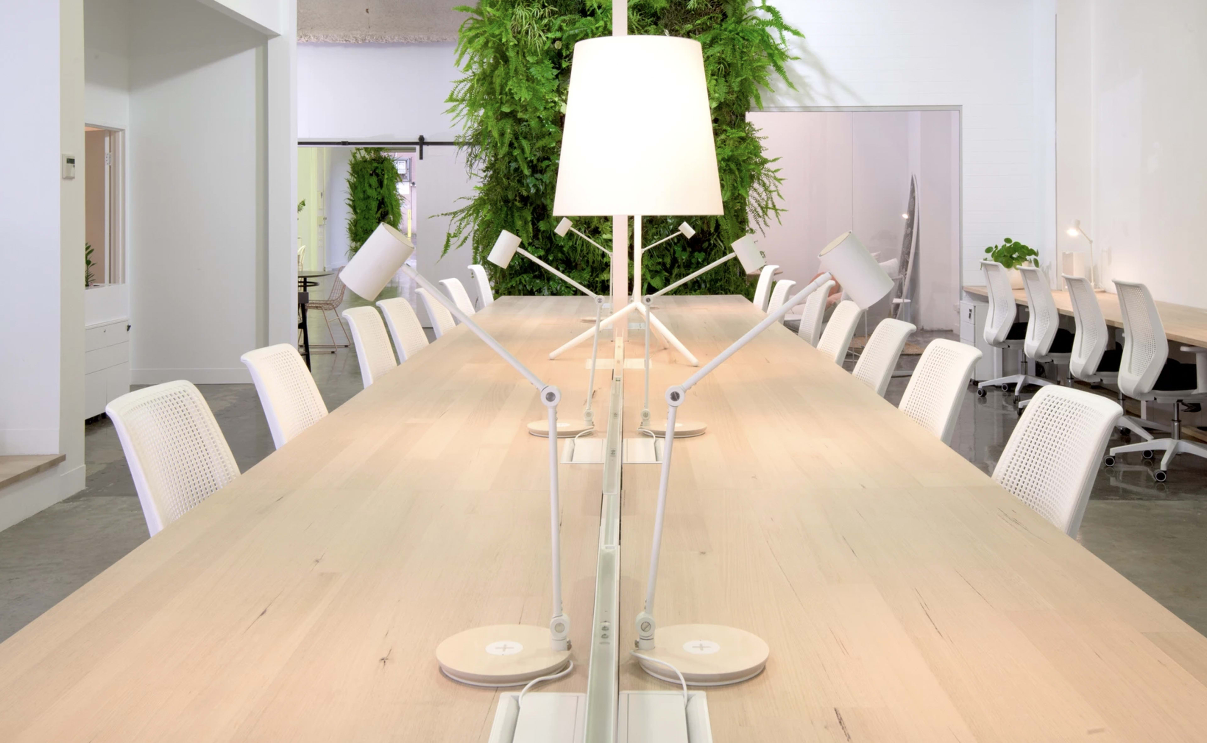 A modern open office space featuring a long wooden table with white chairs, illuminated by a lamp in the center, and greenery in the background.