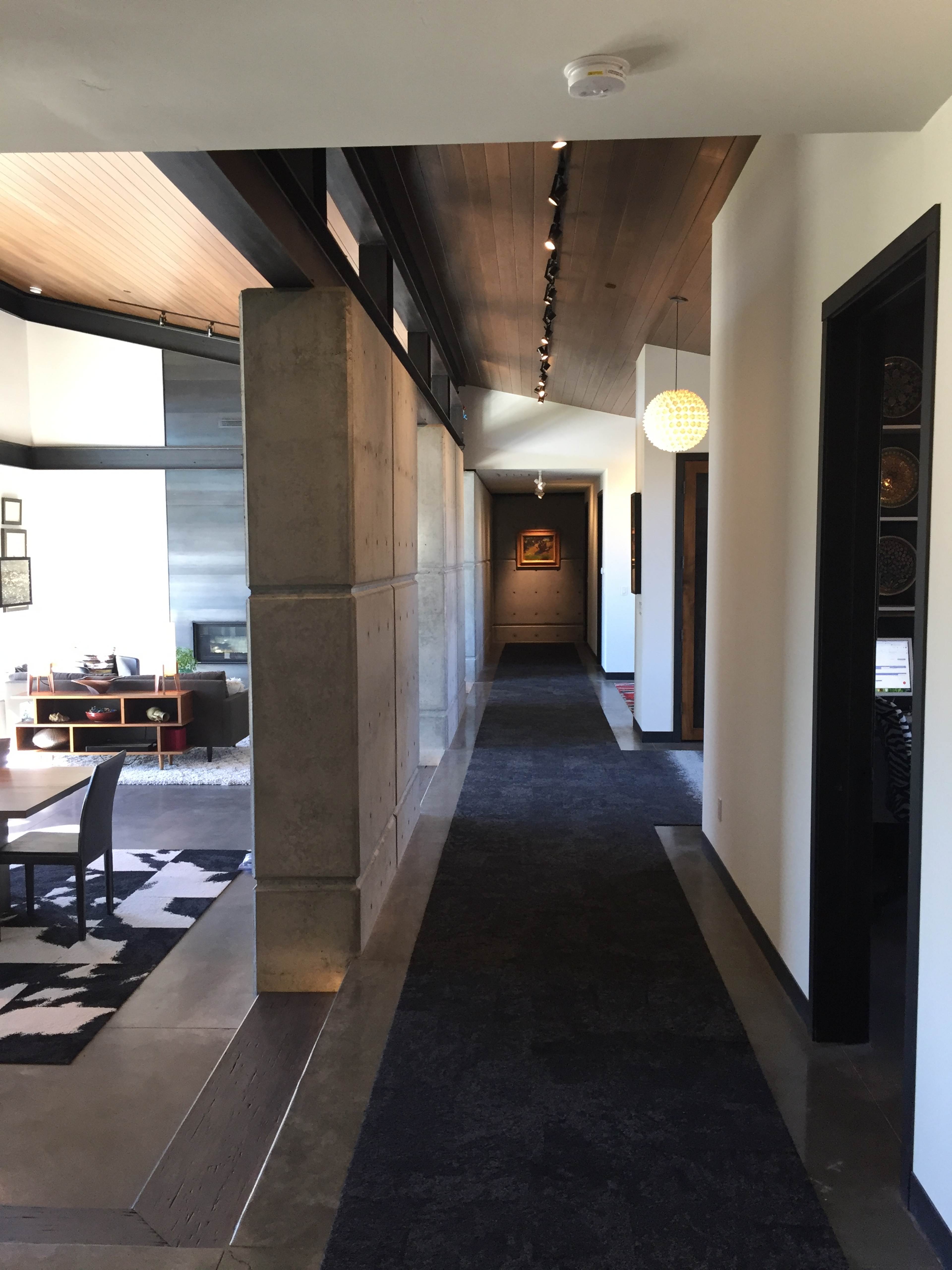 A modern hallway with concrete walls, a dark carpet, and a light fixture hanging from the ceiling, leading to a living area.