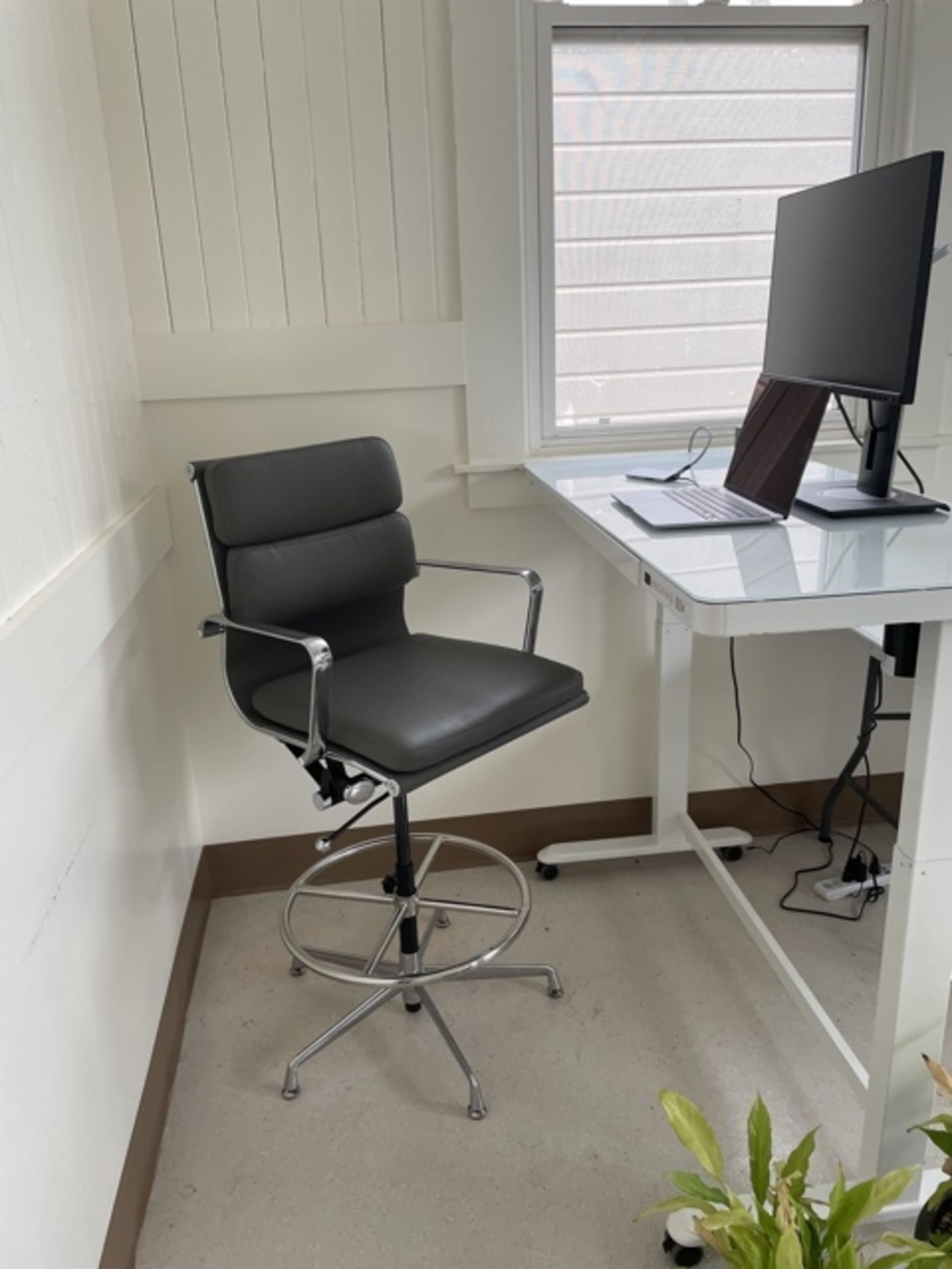 A black bar stool is positioned next to a desk with a computer monitor in a bright room.