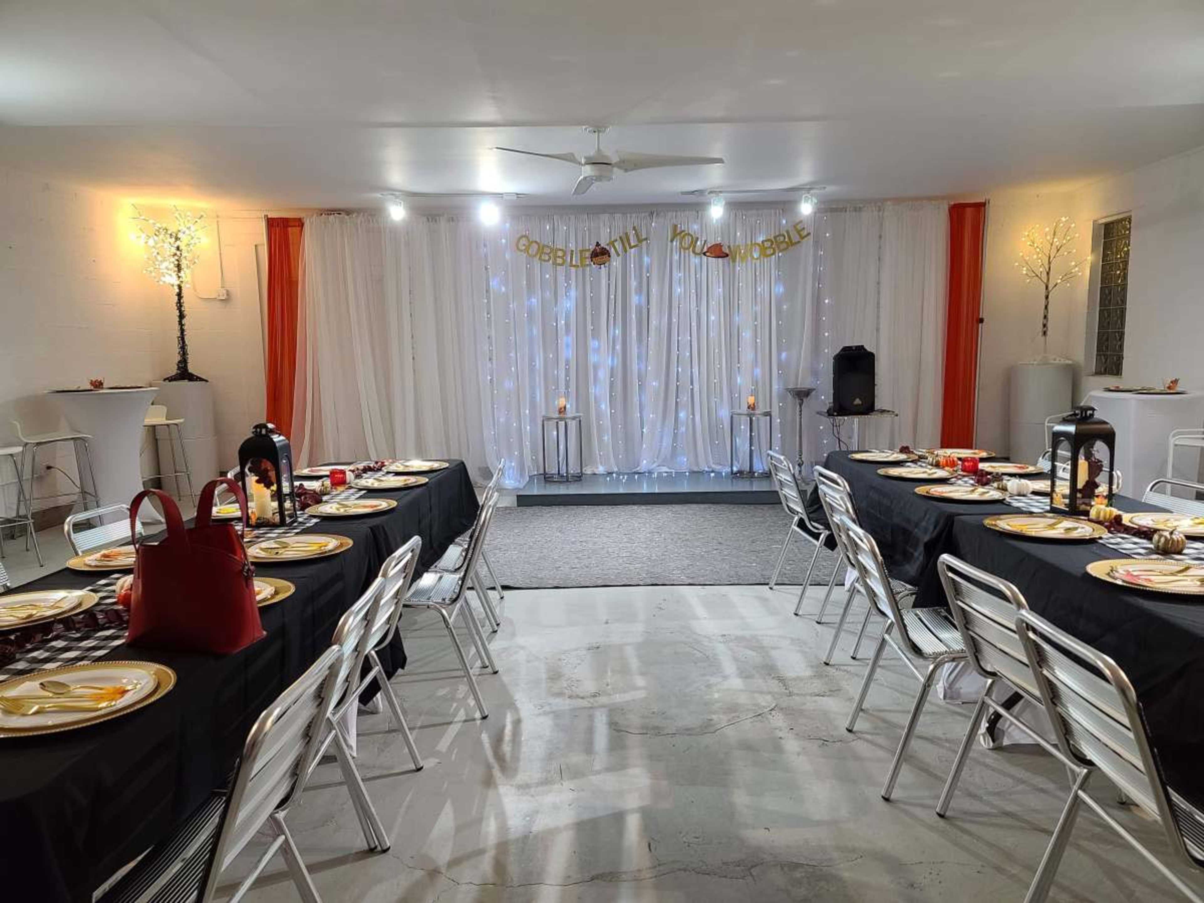 A dining area is set up with black tablecloths and gold plates, illuminated by festive lighting and decorative elements for a gathering.