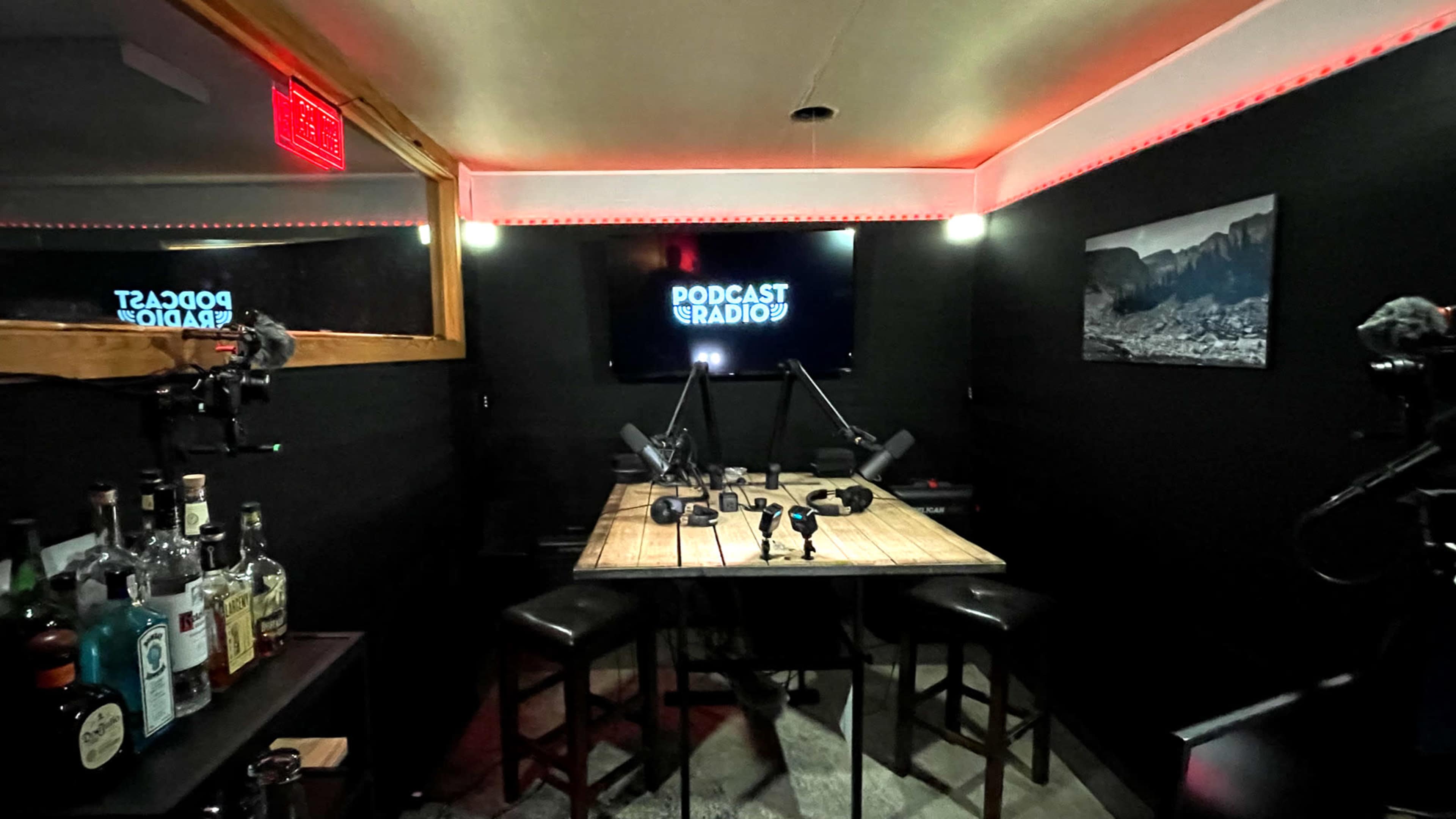 A dimly lit podcast studio with a wooden table, microphones, and a backlit sign reading "PODCAST RADIO."