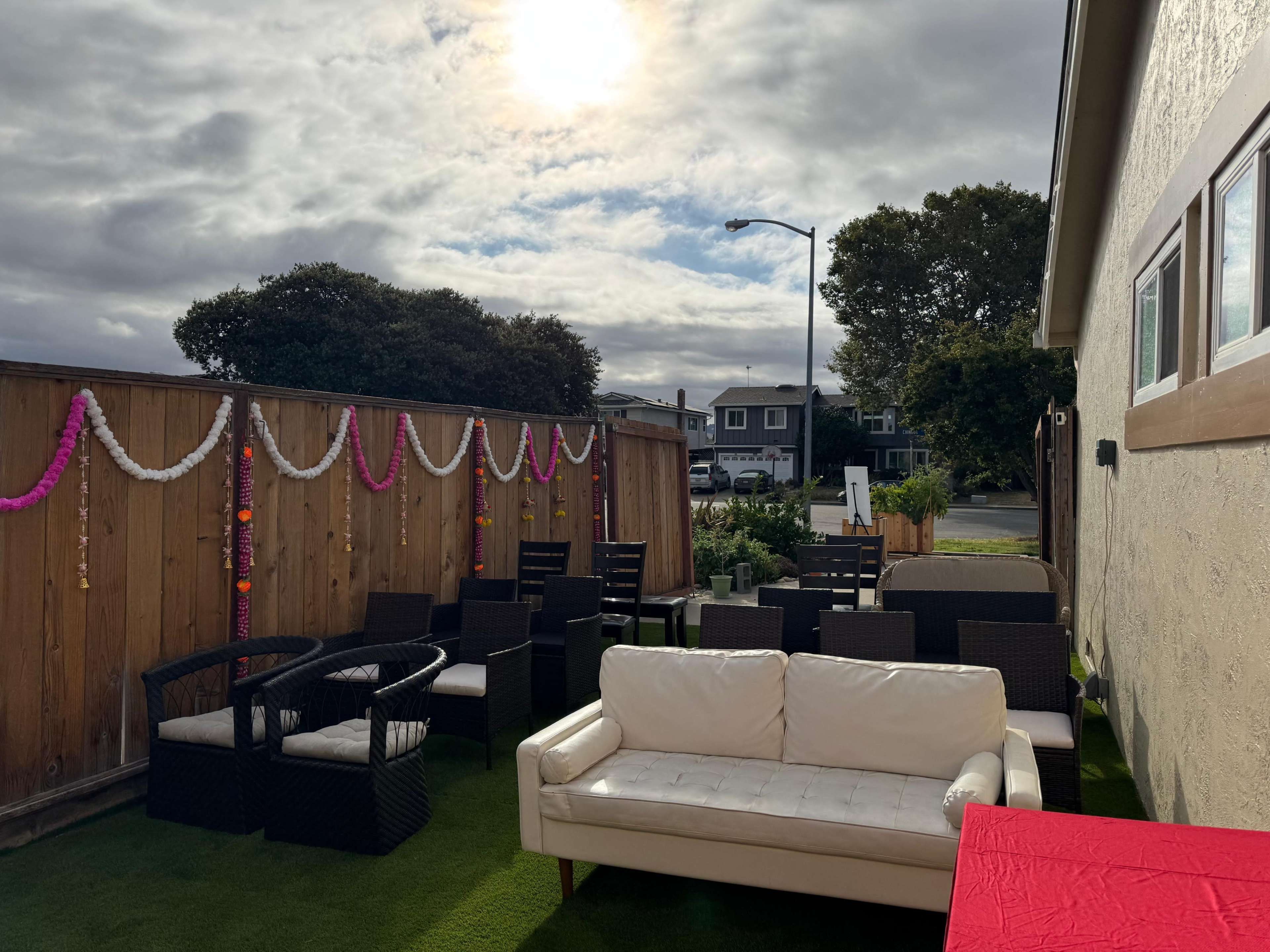 A patio area with a white couch and several wicker chairs arranged on green grass, framed by a wooden fence decorated with colorful garlands, under a partly cloudy sky.