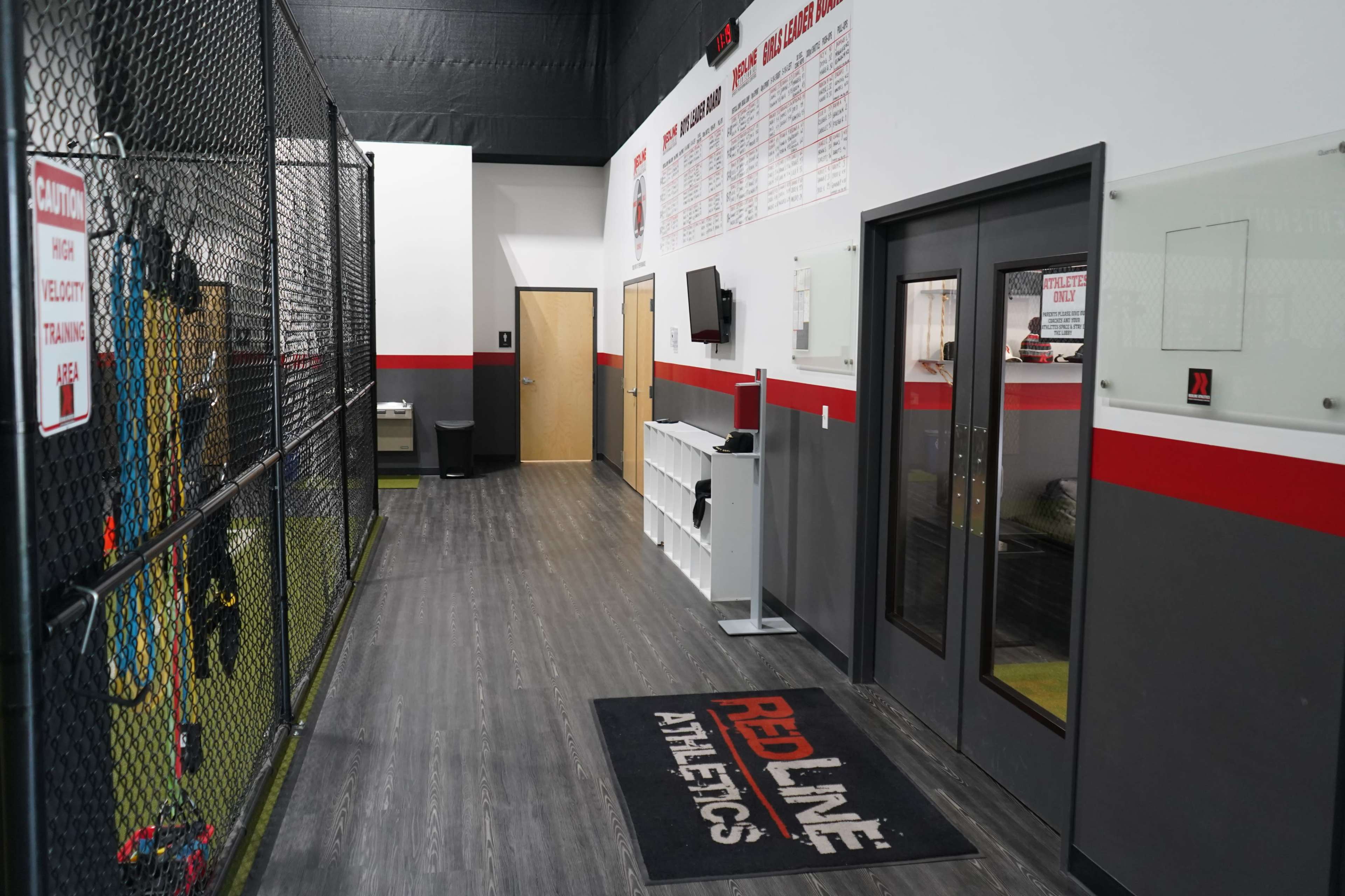 The image shows a sports training facility hallway featuring a black mat with "RED LINE ATHLETICS" logo, white and red walls, and a viewing window into a training area behind a black fence.