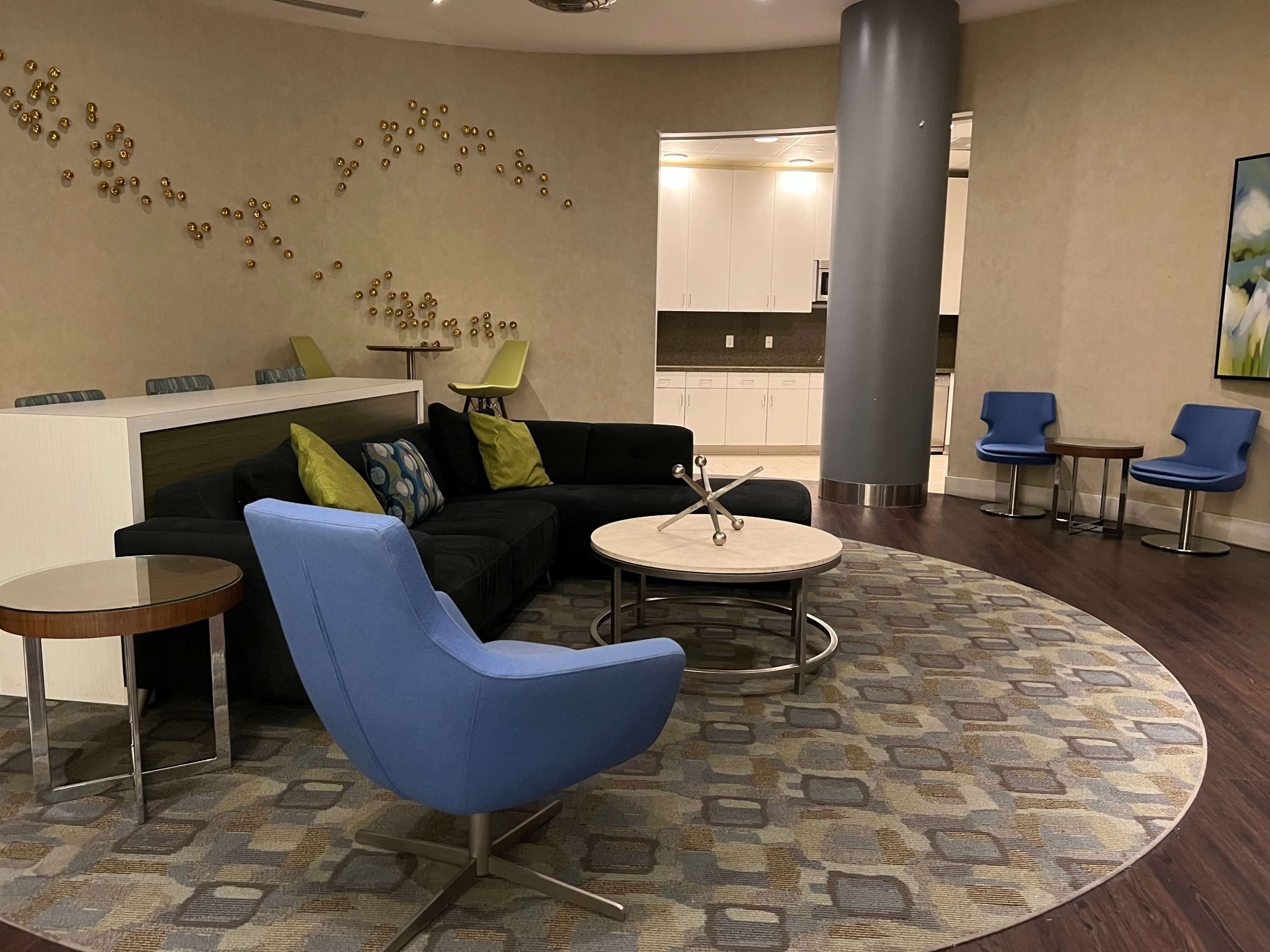 The scene shows a modern lounge area with a dark sofa, a circular coffee table, two blue chairs, and a patterned rug, complemented by a kitchenette in the background.