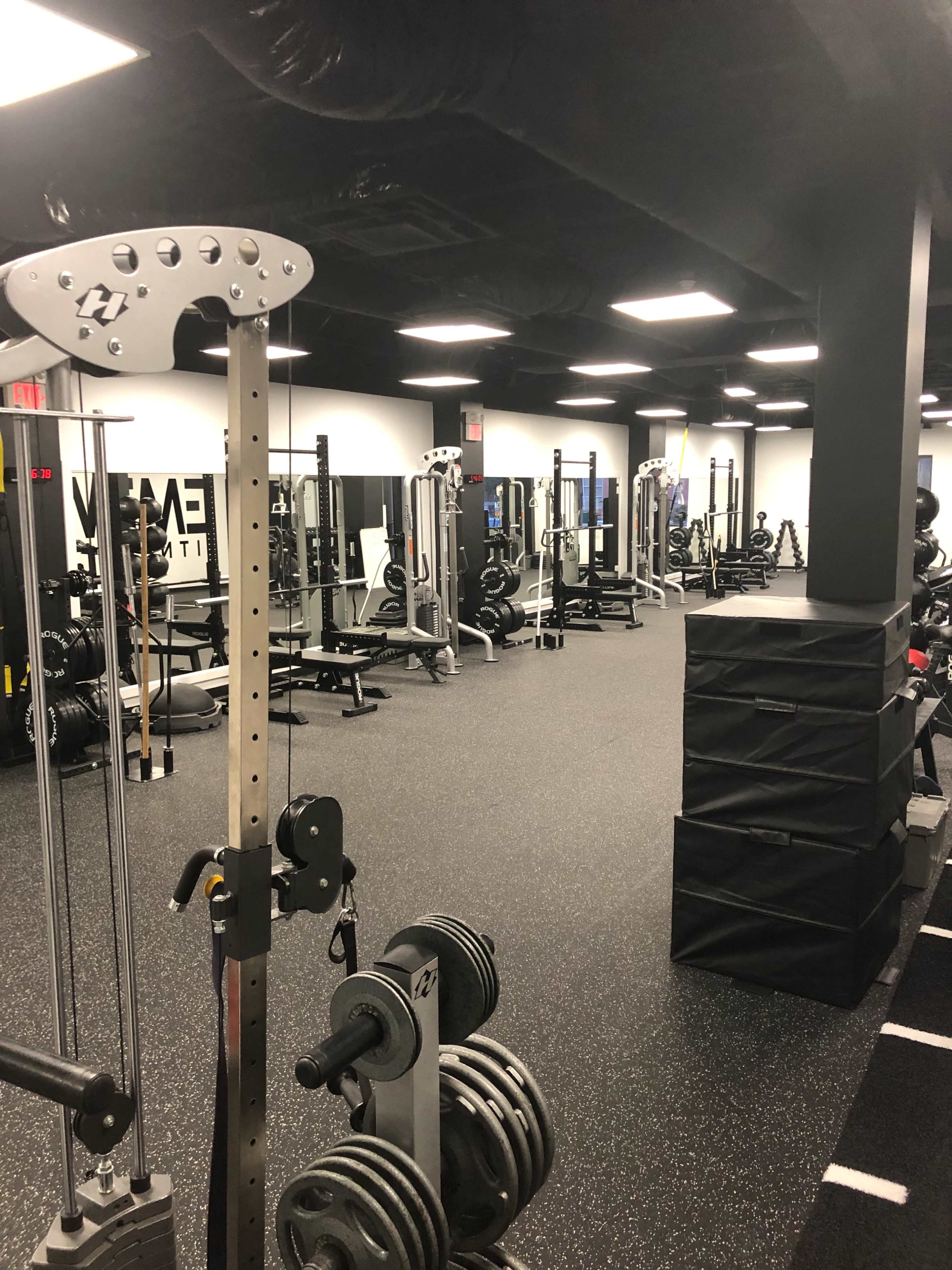 The image shows an indoor gym facility with various weightlifting equipment and a black rubberized floor.