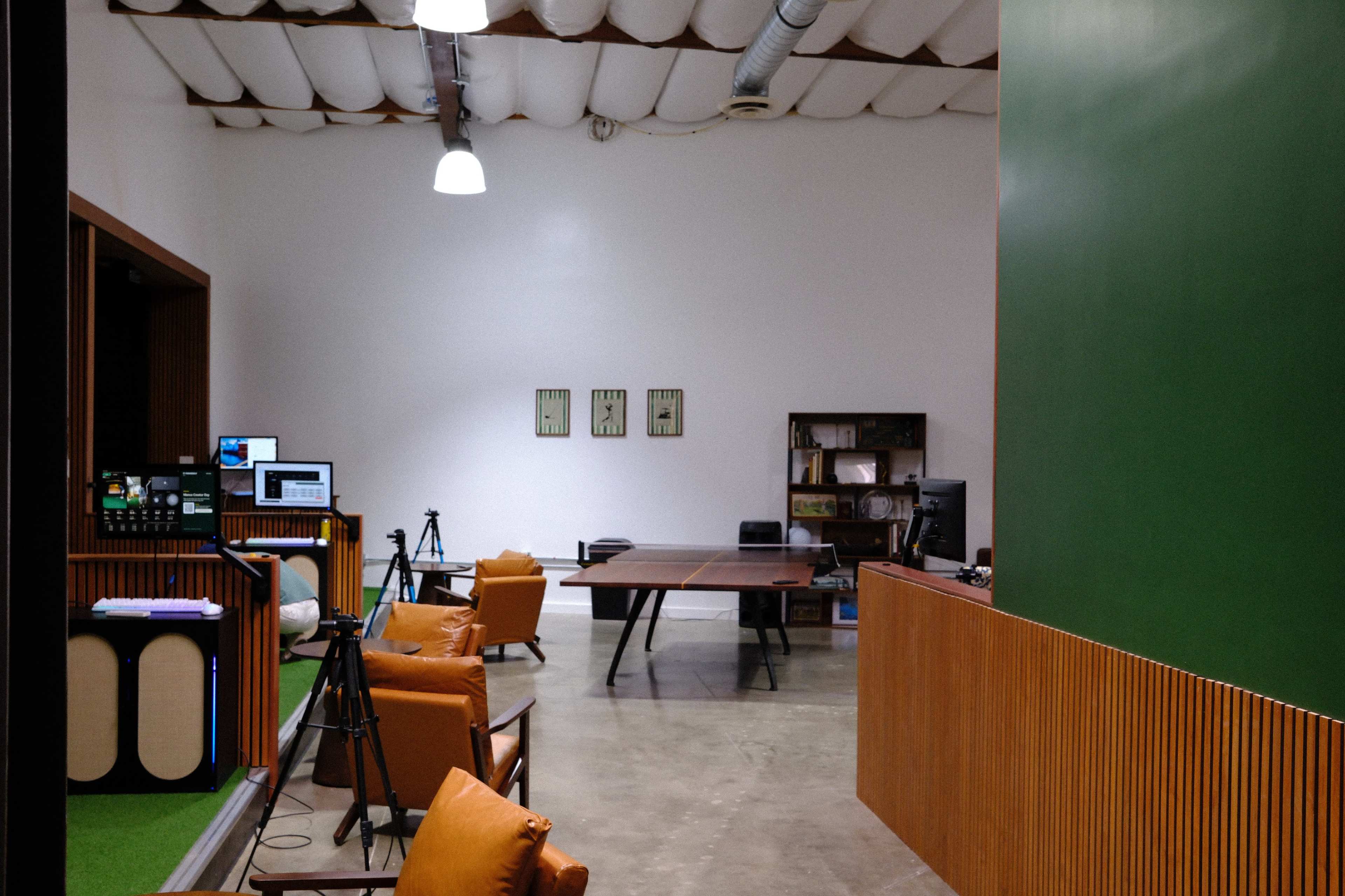 The image shows a modern office space featuring a large wooden table, several cameras set up for recording, and brown leather chairs.