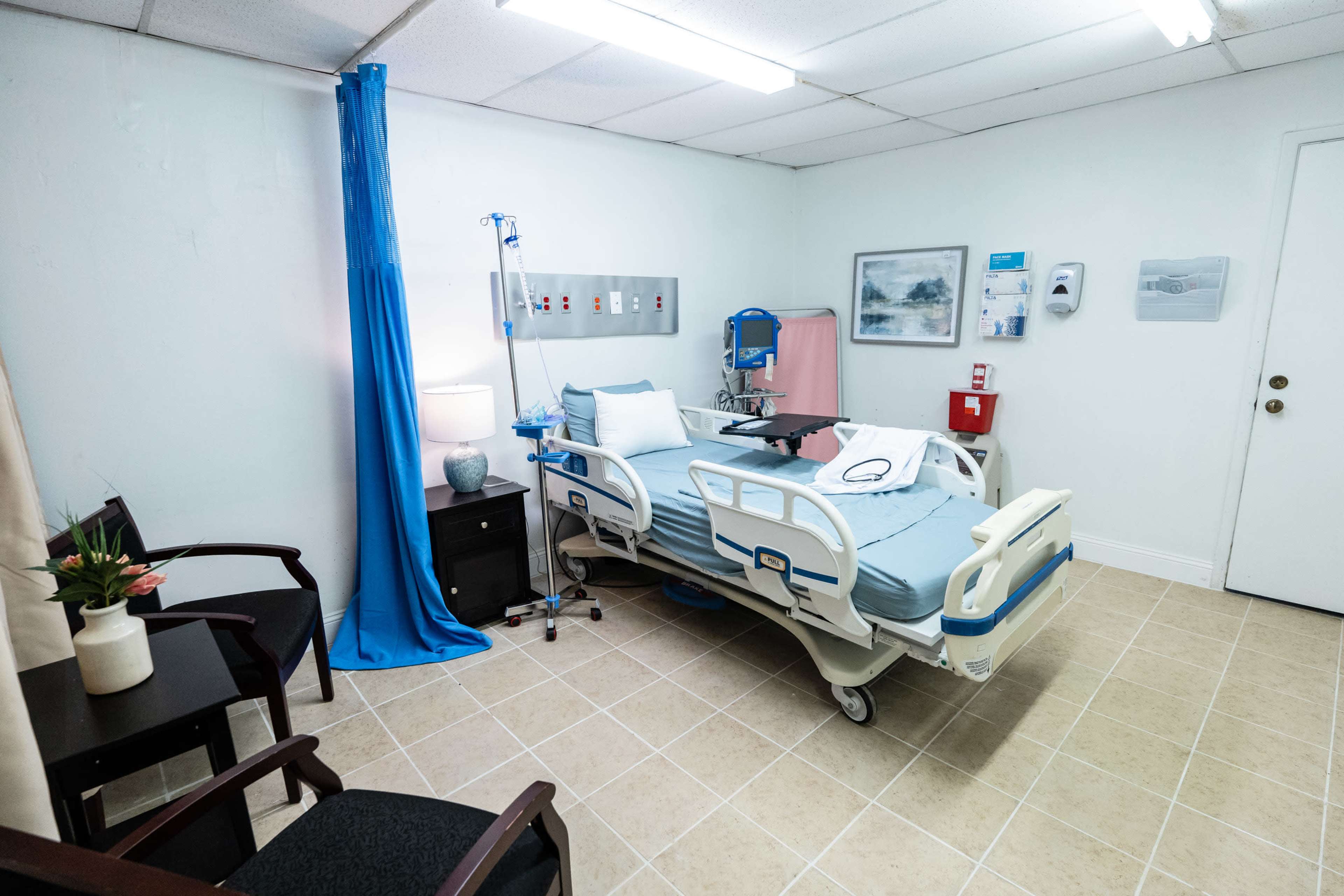 The image shows a hospital room featuring a patient bed, medical equipment, a small bedside table, and a chair in a tidy environment.