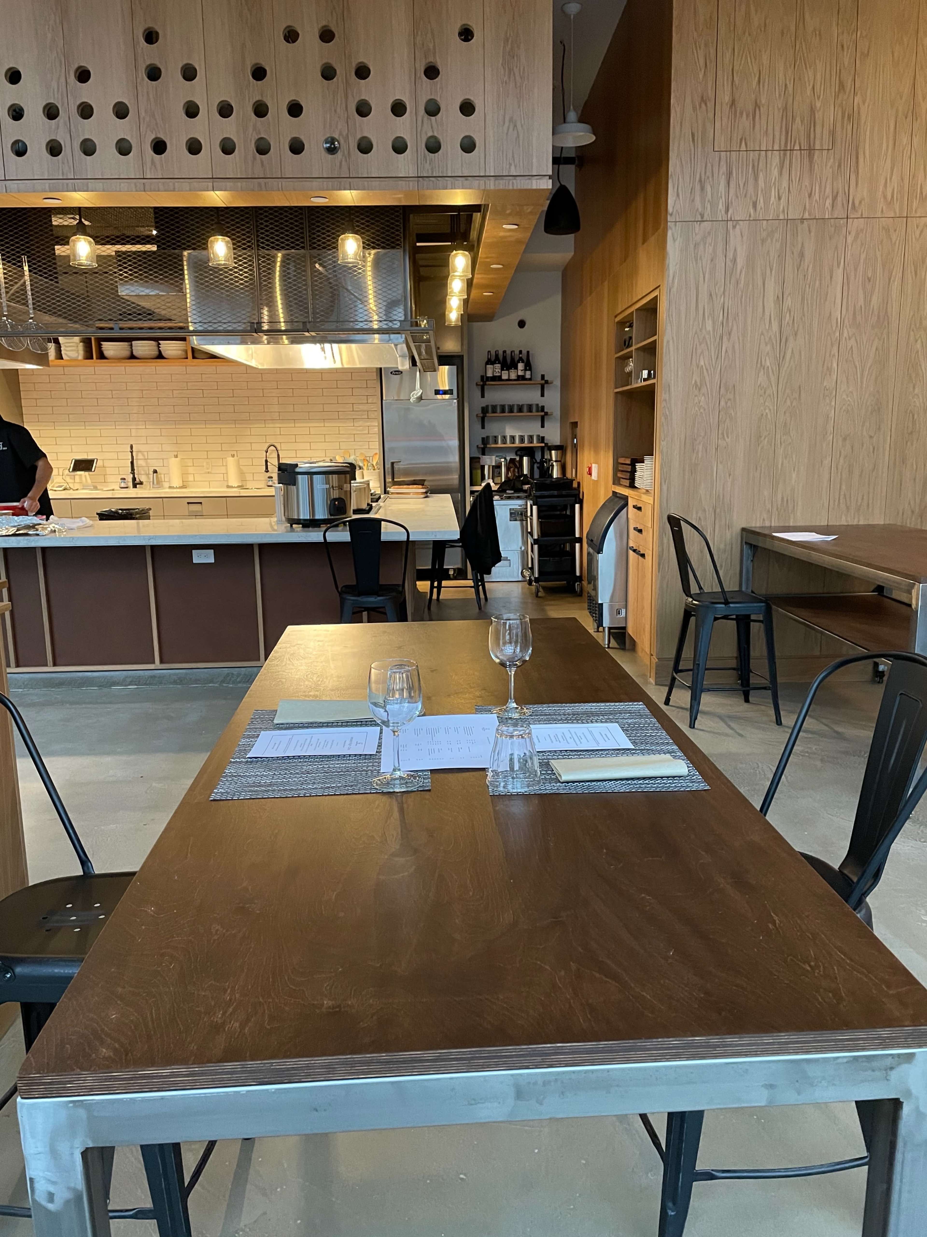 A wooden dining table is set for two with menus and glasses, next to a modern kitchen with stainless steel appliances and wooden cabinetry.