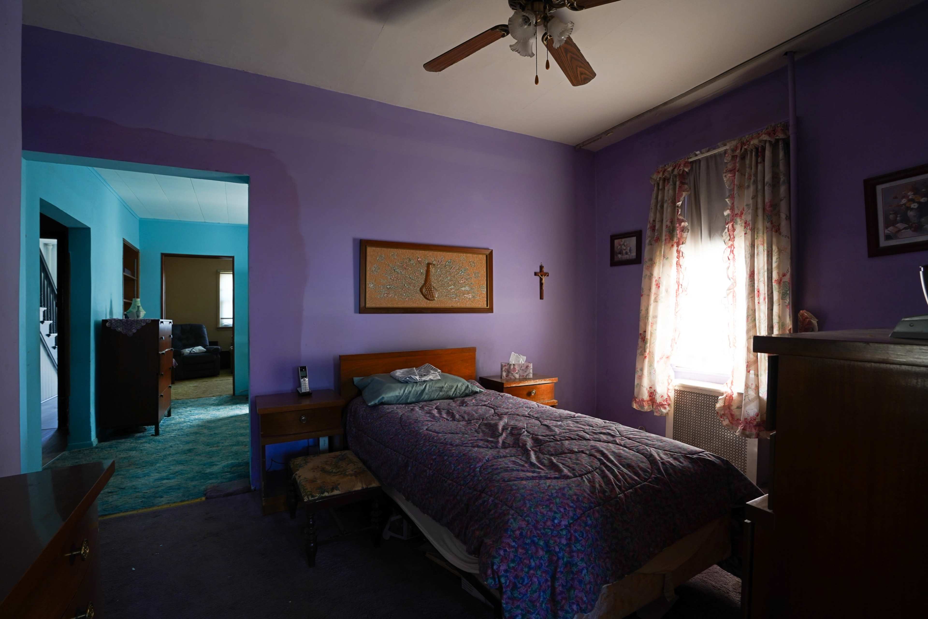 A small bedroom with purple walls, a bed, and a window letting in natural light, is connected to a blue-tinted hallway.