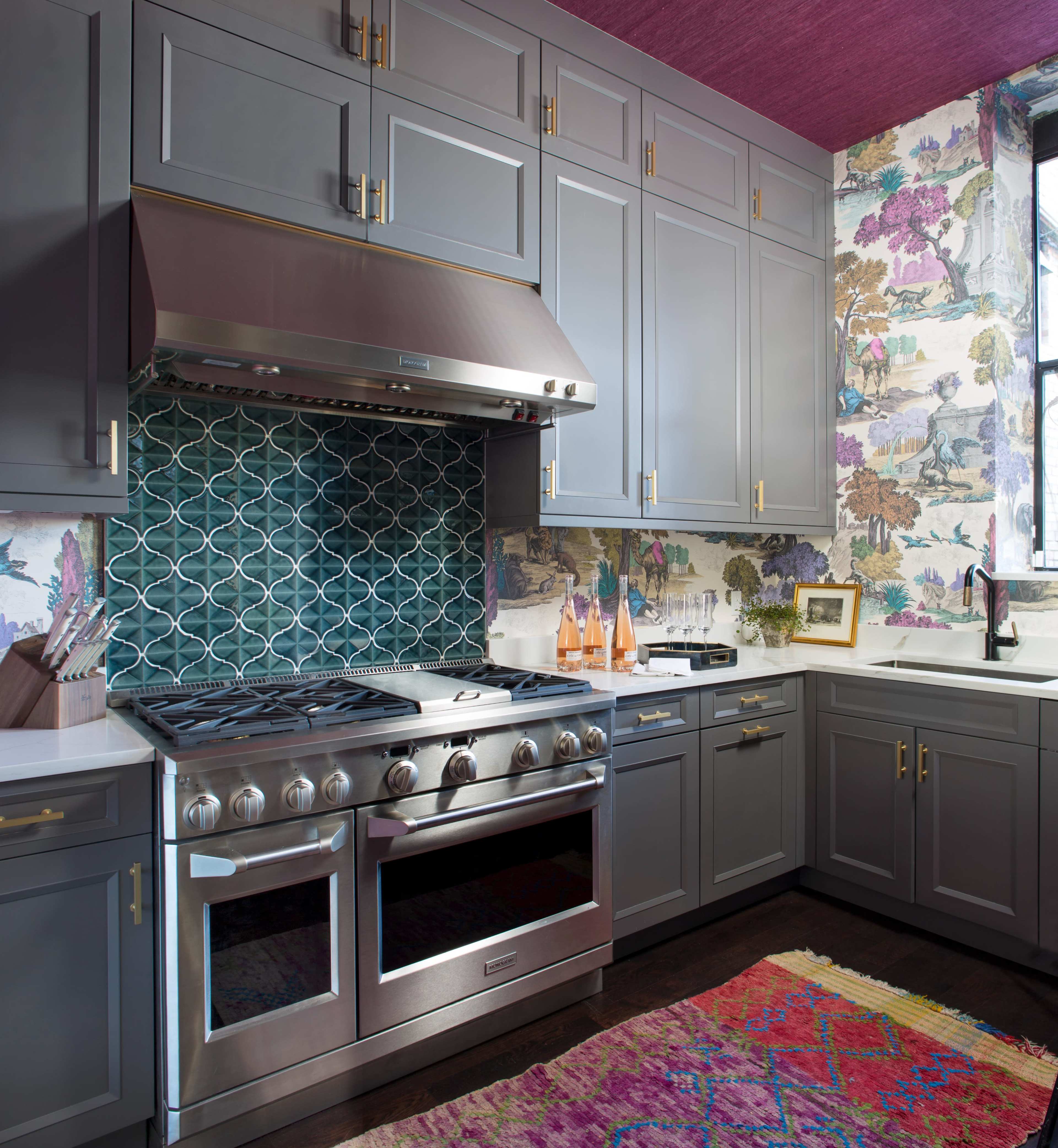 The image shows a contemporary kitchen with gray cabinets, a stainless steel range hood, and a colorful tiled backsplash beneath a window with floral wallpaper.