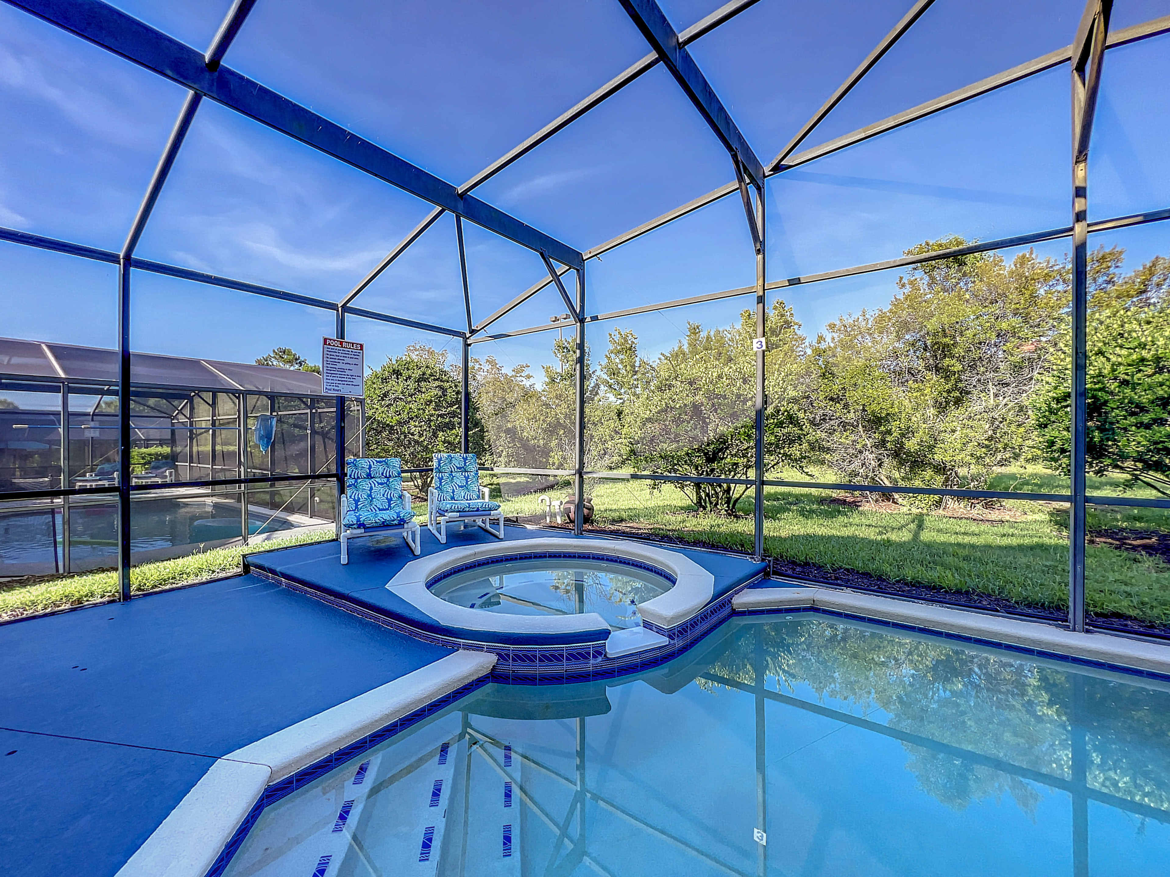 The image shows a screened-in pool area with a hot tub and two lounge chairs, surrounded by greenery and a clear blue sky.
