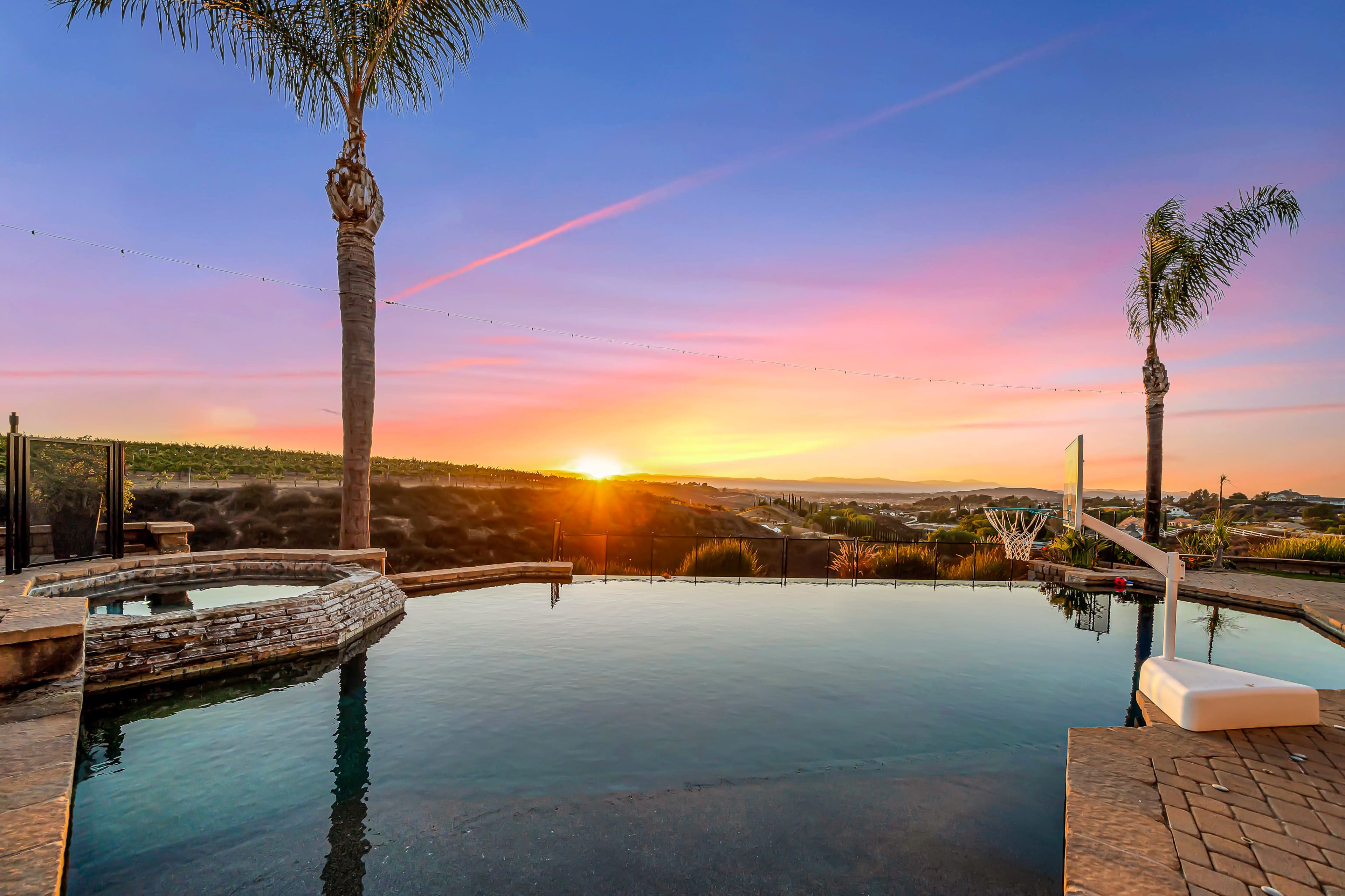 The image shows a swimming pool with palm trees at sunset, reflecting vibrant colors in the water, overlooking a hilly landscape.