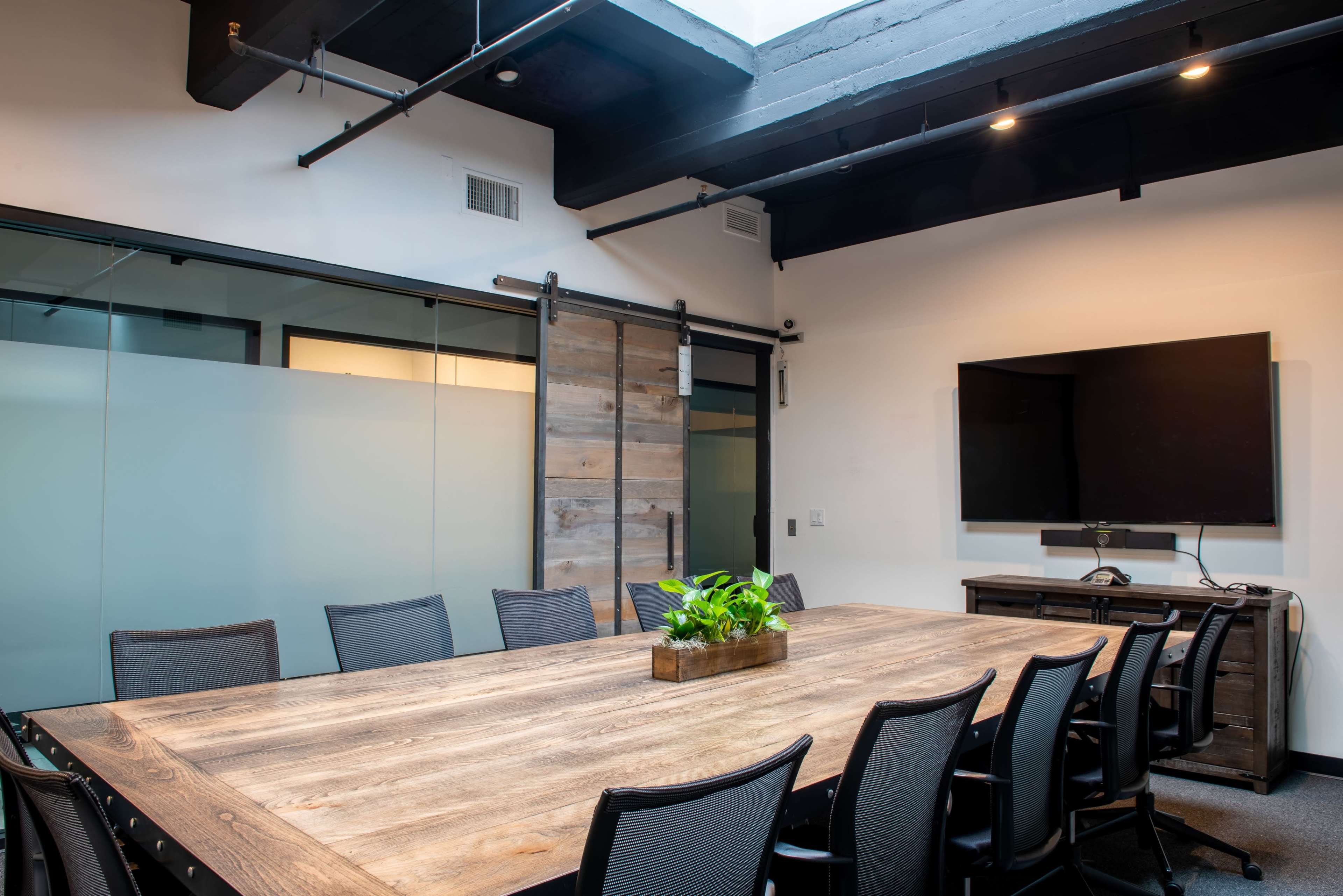 A modern conference room with a large wooden table, black office chairs, and a wall-mounted screen.