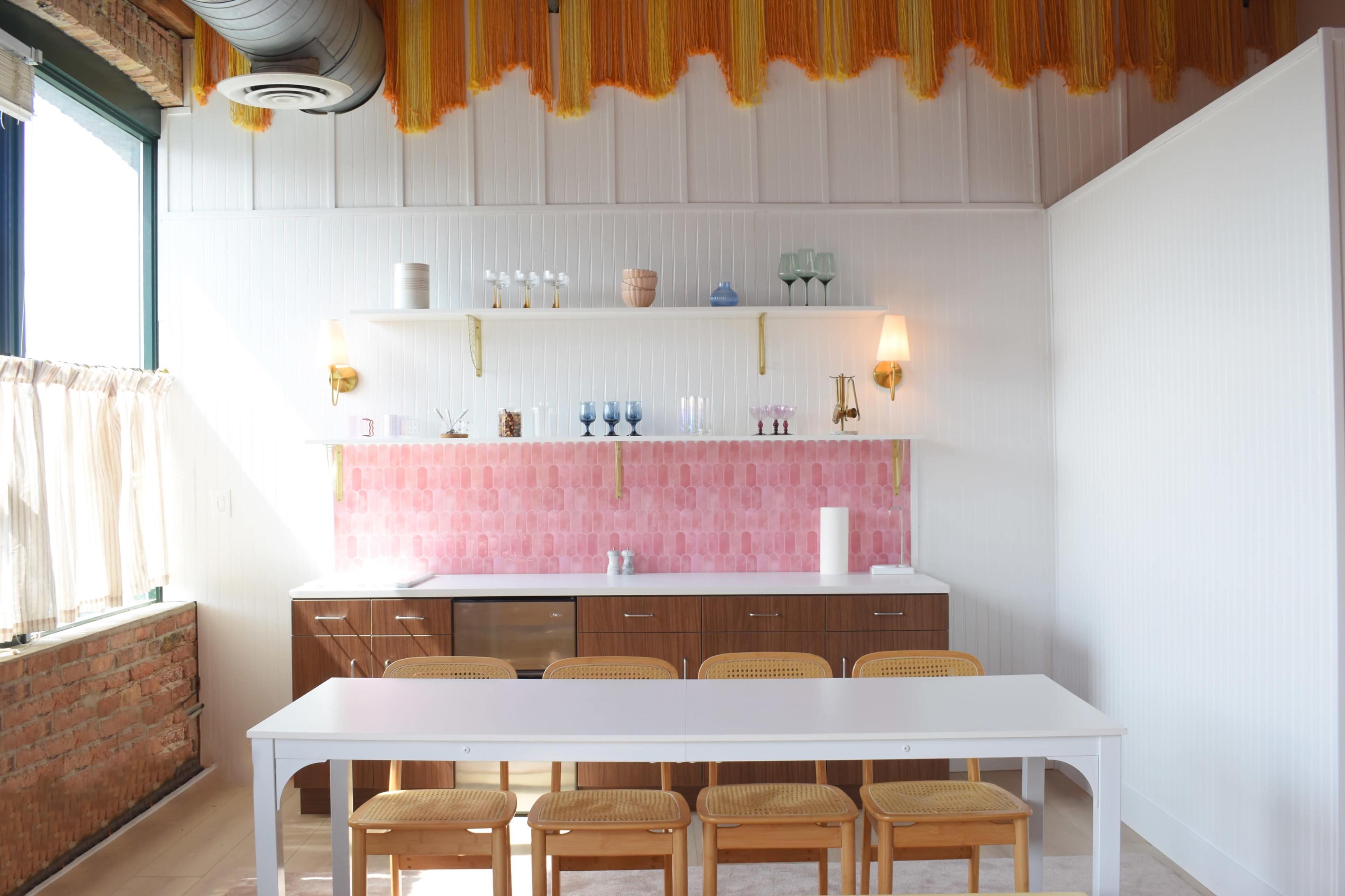 The image shows a modern interior space featuring a kitchen area with a pink tiled backsplash, wooden cabinetry, a long white dining table, and rattan chairs.