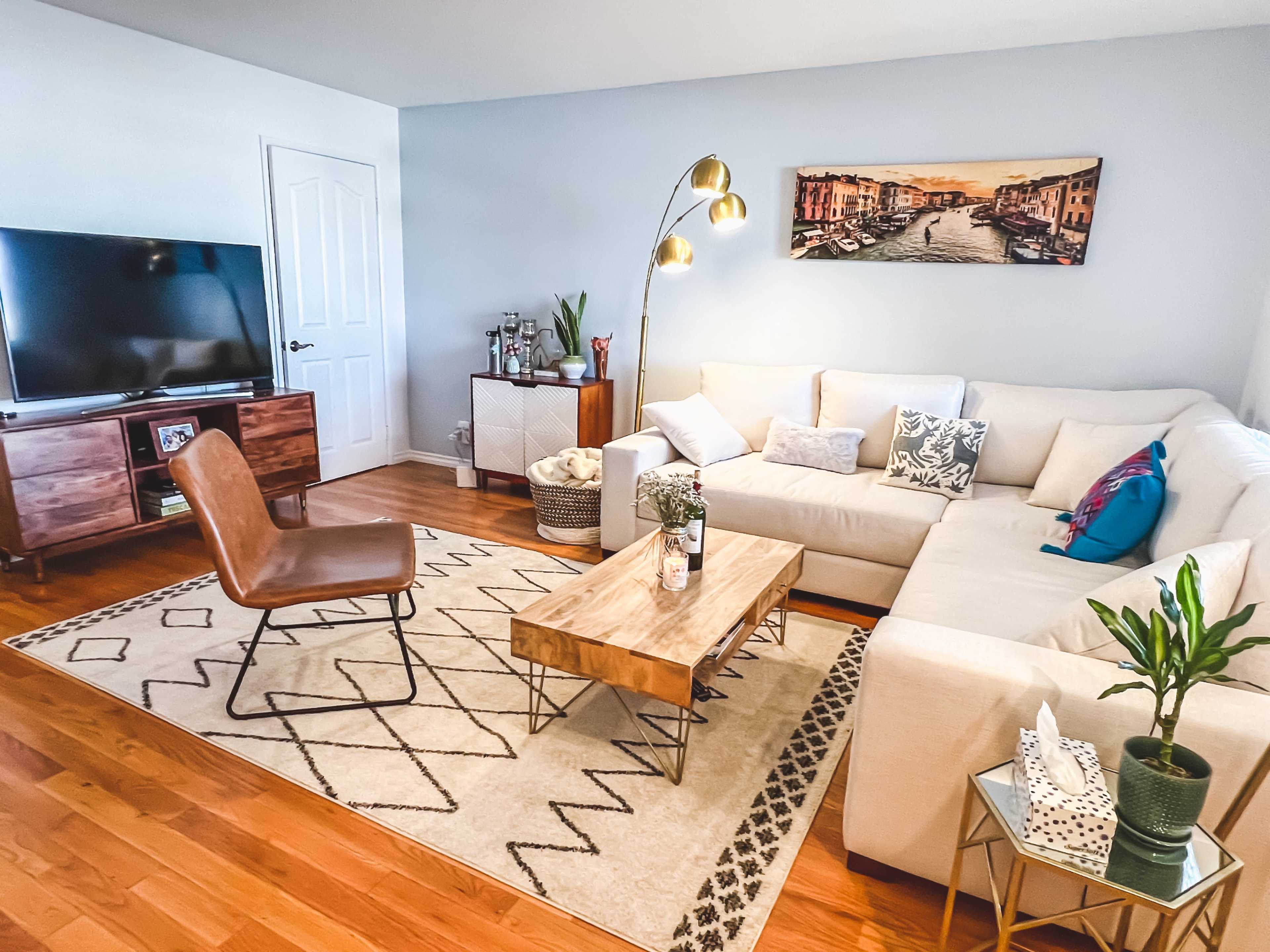 The image shows a modern living room featuring a light-colored sectional sofa, a wooden coffee table, a decorative area rug, and a television on a wooden stand.