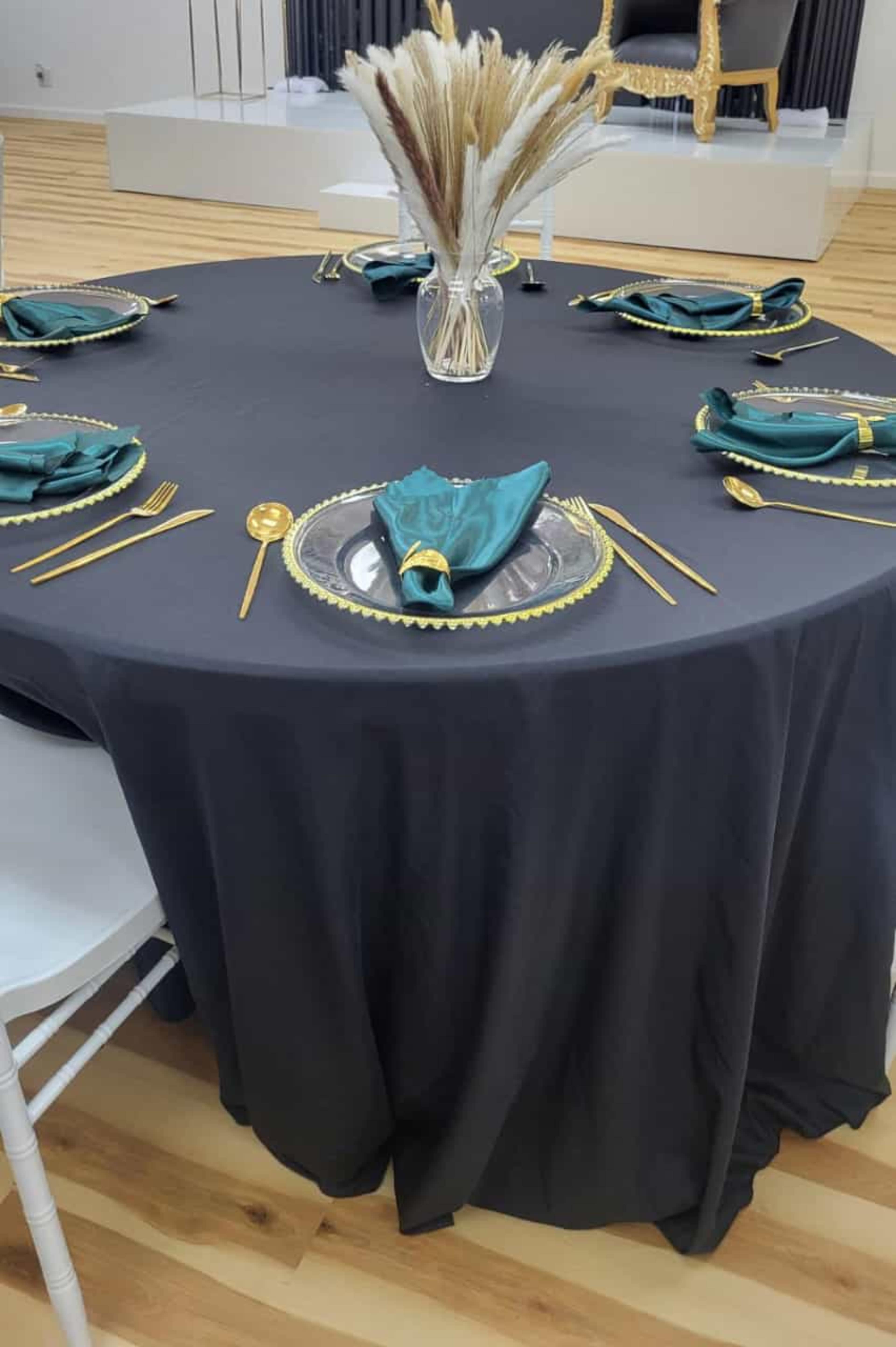 A round black table is set with gold-trimmed plates, teal napkins, and a vase of dried flowers in the center.