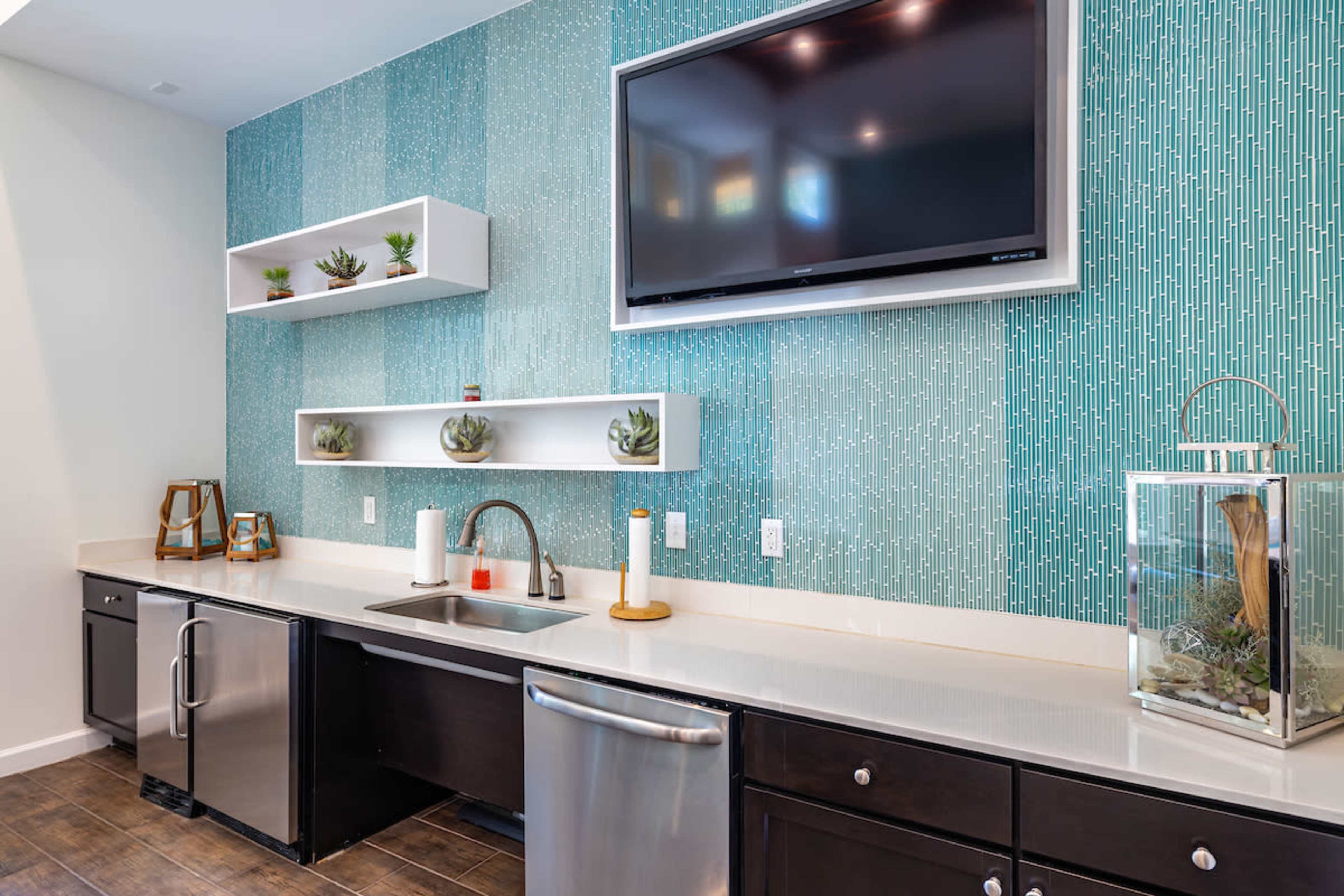 A modern kitchen displays a turquoise patterned wall, a stainless steel sink, a television mounted above the counter, and decorative shelves with plants.
