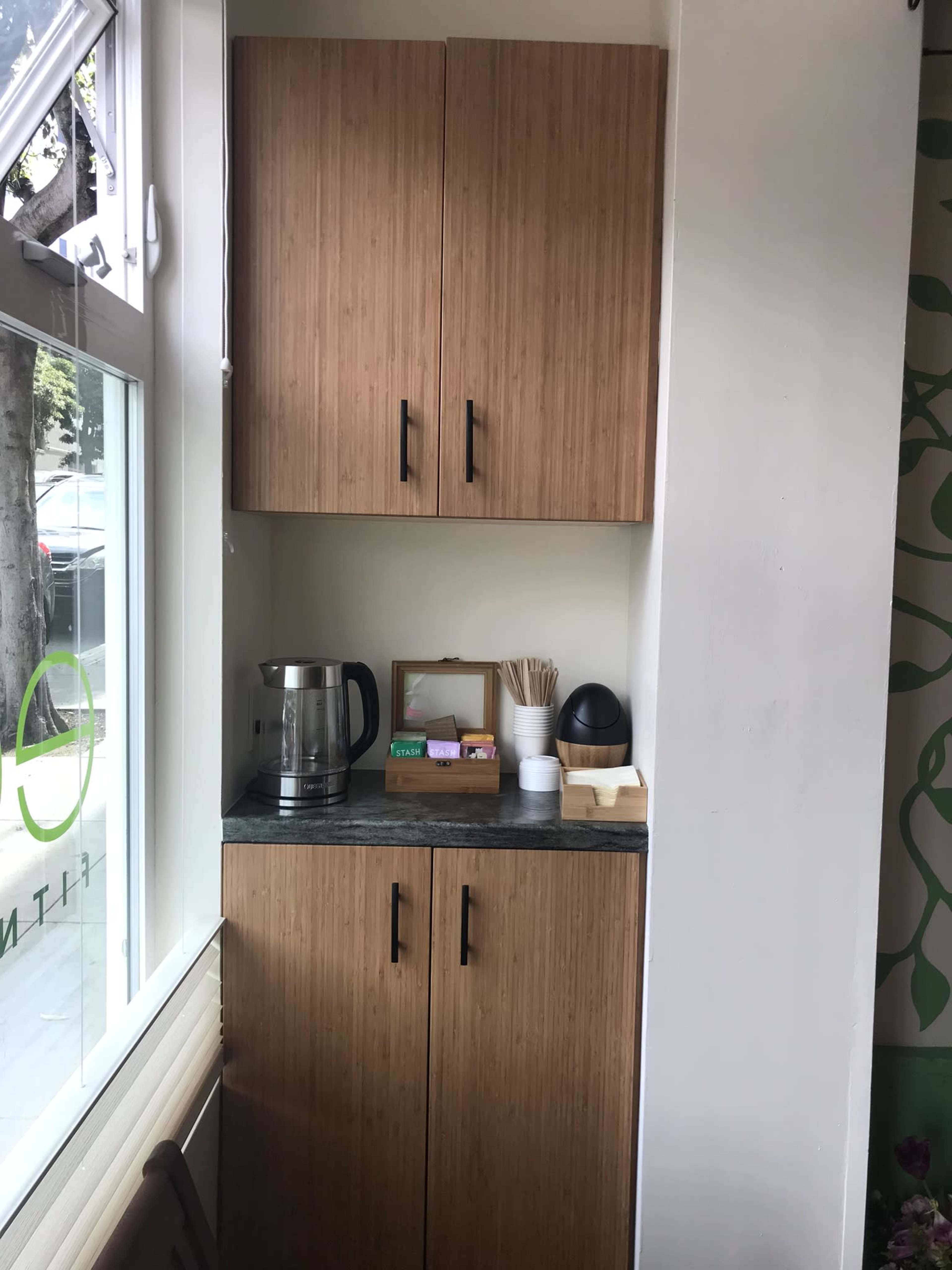 The image shows a corner of a café with a wooden cabinet featuring two doors, a countertop with a kettle, utensils, and a small framed display.