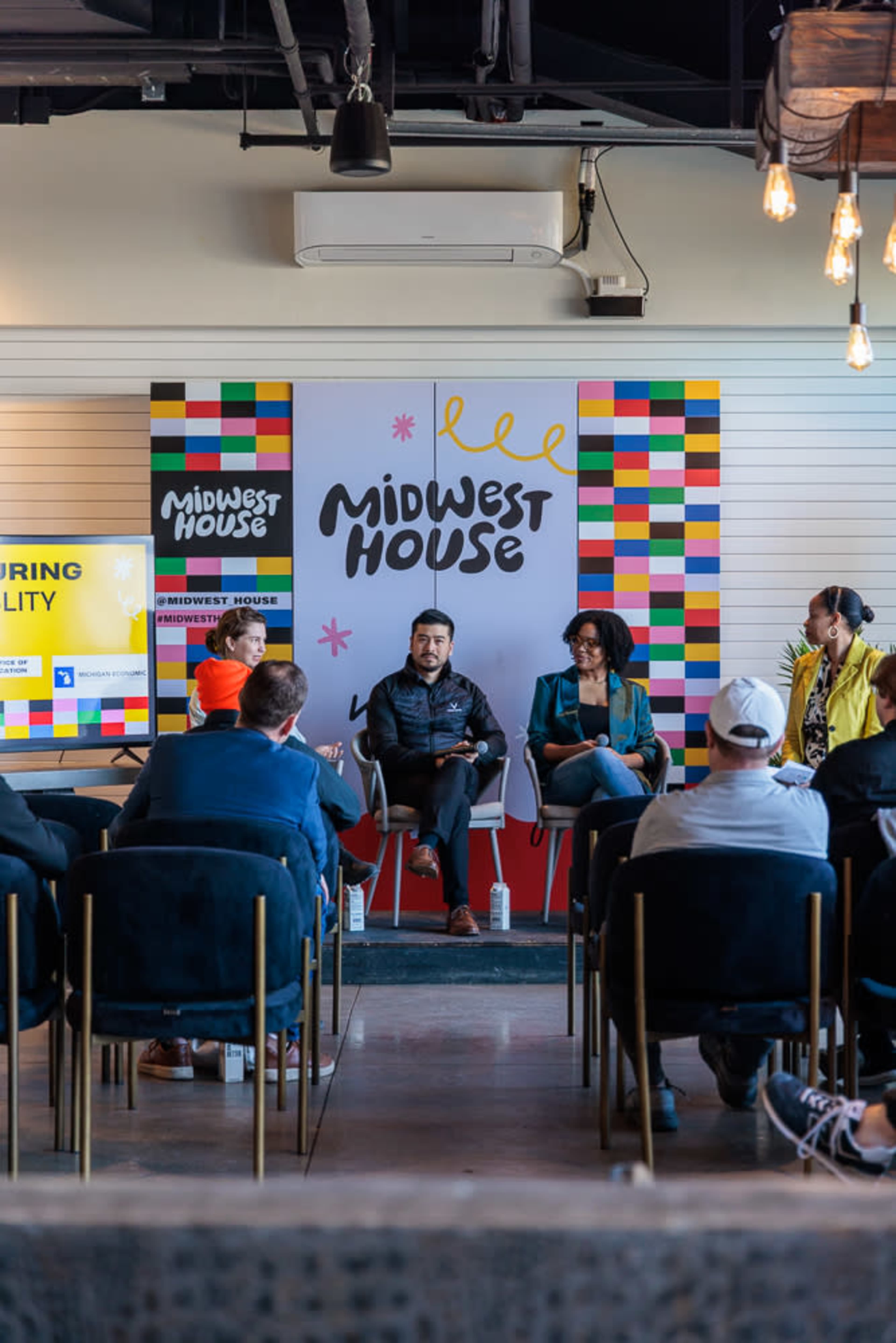 A panel discussion is taking place at Midwest House, featuring two speakers on stage and an audience seated in rows.