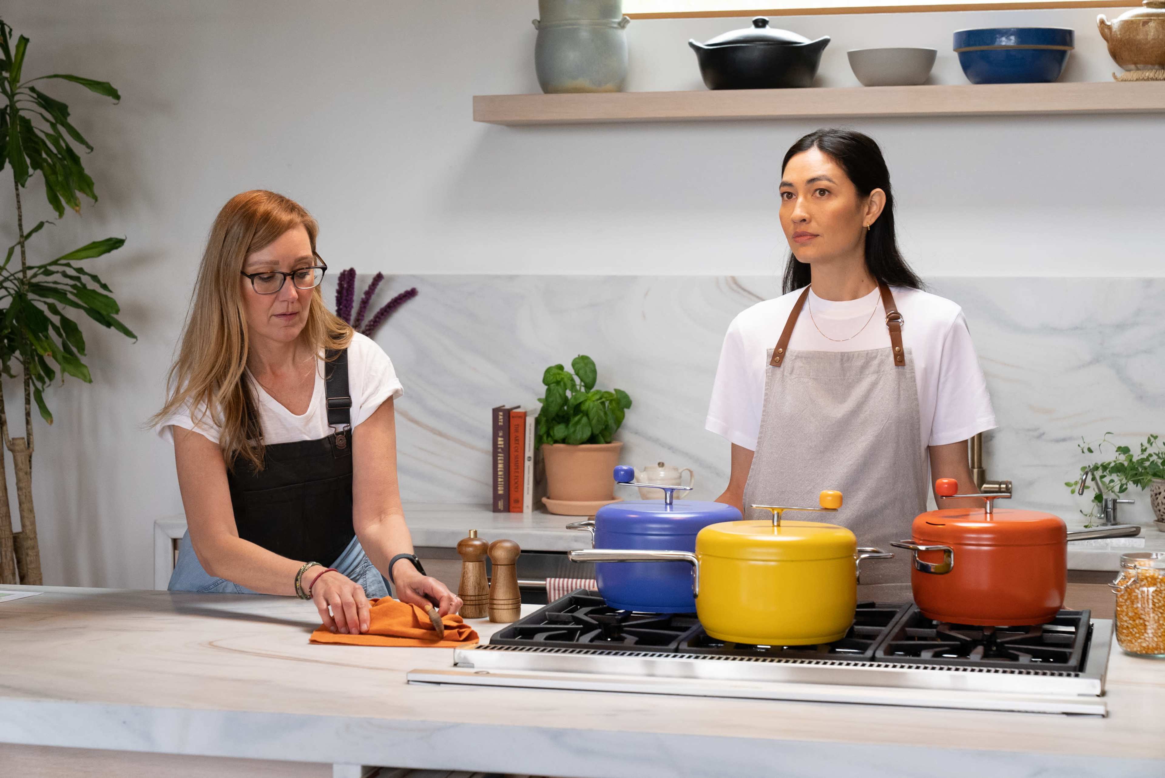 Two women stand in a modern kitchen with colorful cooking pots on the stove and various kitchen items neatly arranged on the counter.