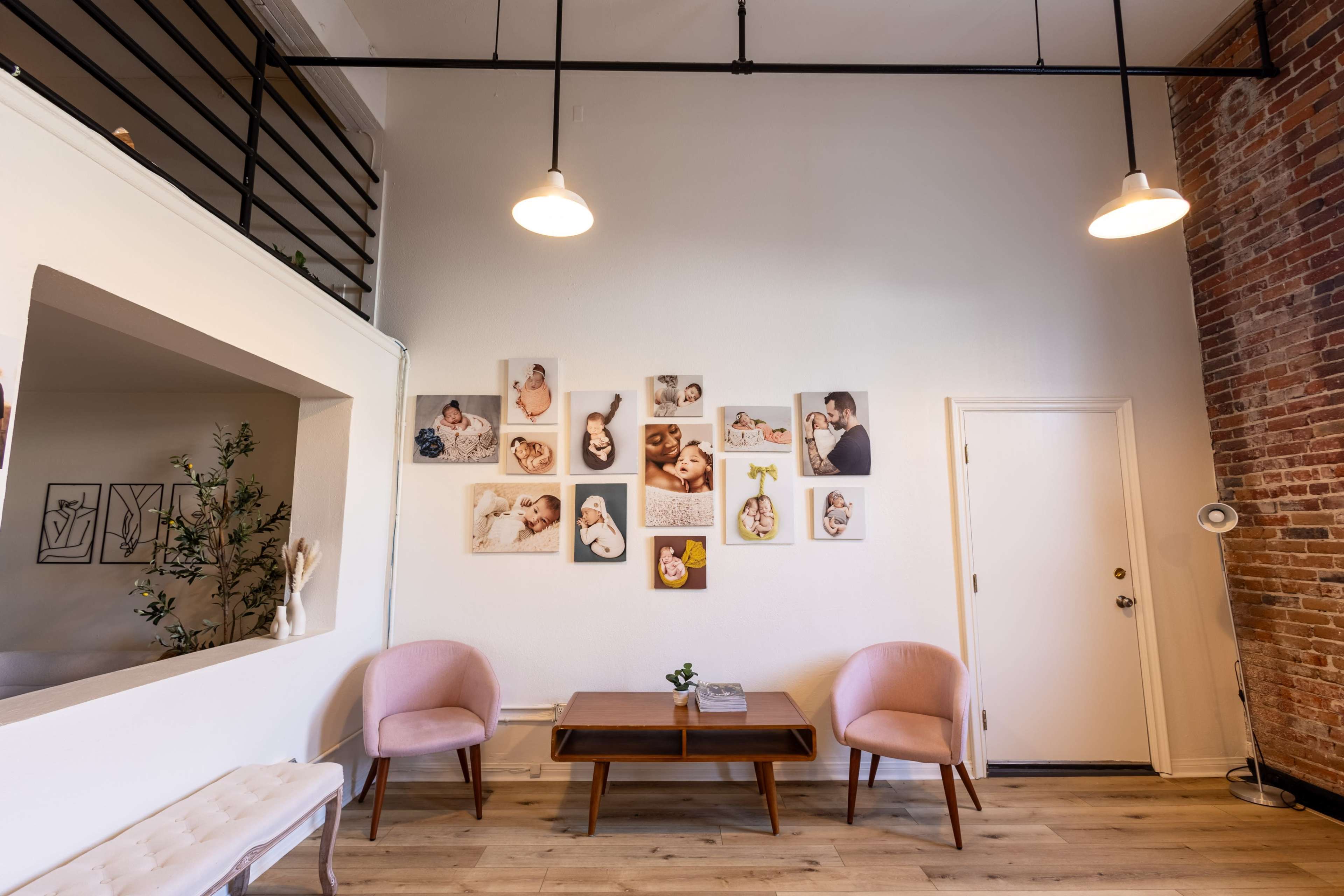 A modern waiting area features two pink chairs, a coffee table, and a gallery wall adorned with framed photographs, alongside exposed brick walls.