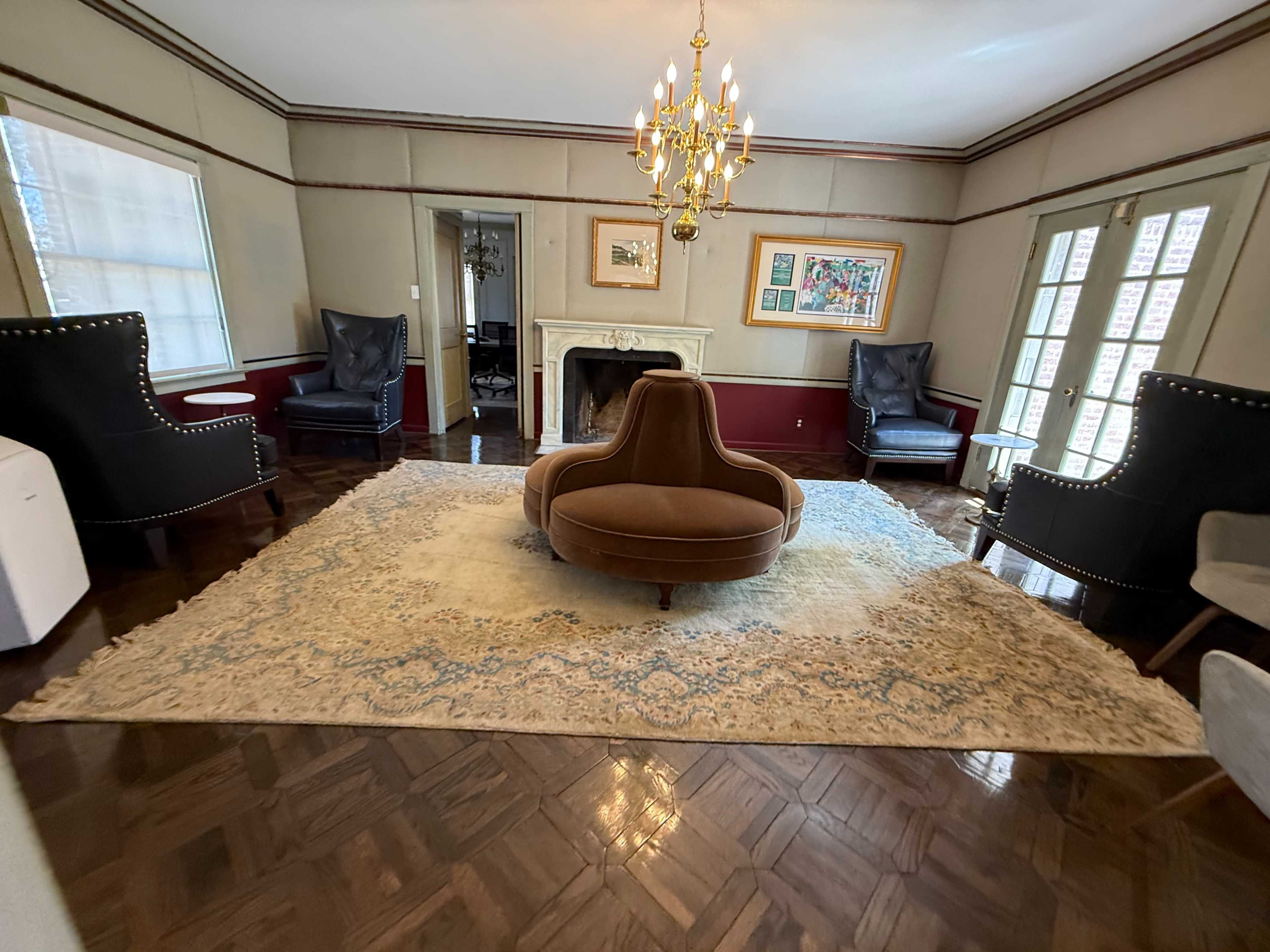 The room features a central circular brown sofa surrounded by four armchairs on a large area rug, with a chandelier hanging from the ceiling and a fireplace in the background.