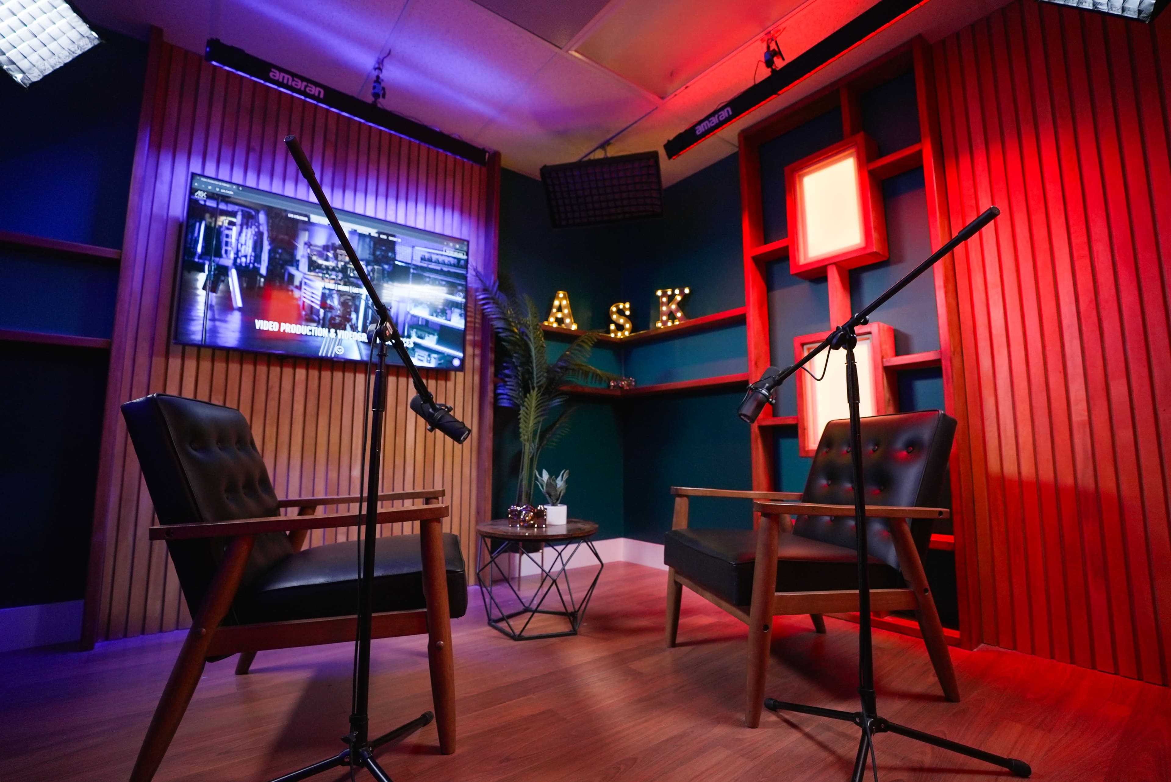 The image shows a small studio setup with two chairs, each equipped with a microphone on a stand, and a brightly lit background featuring wooden panels and a television screen.