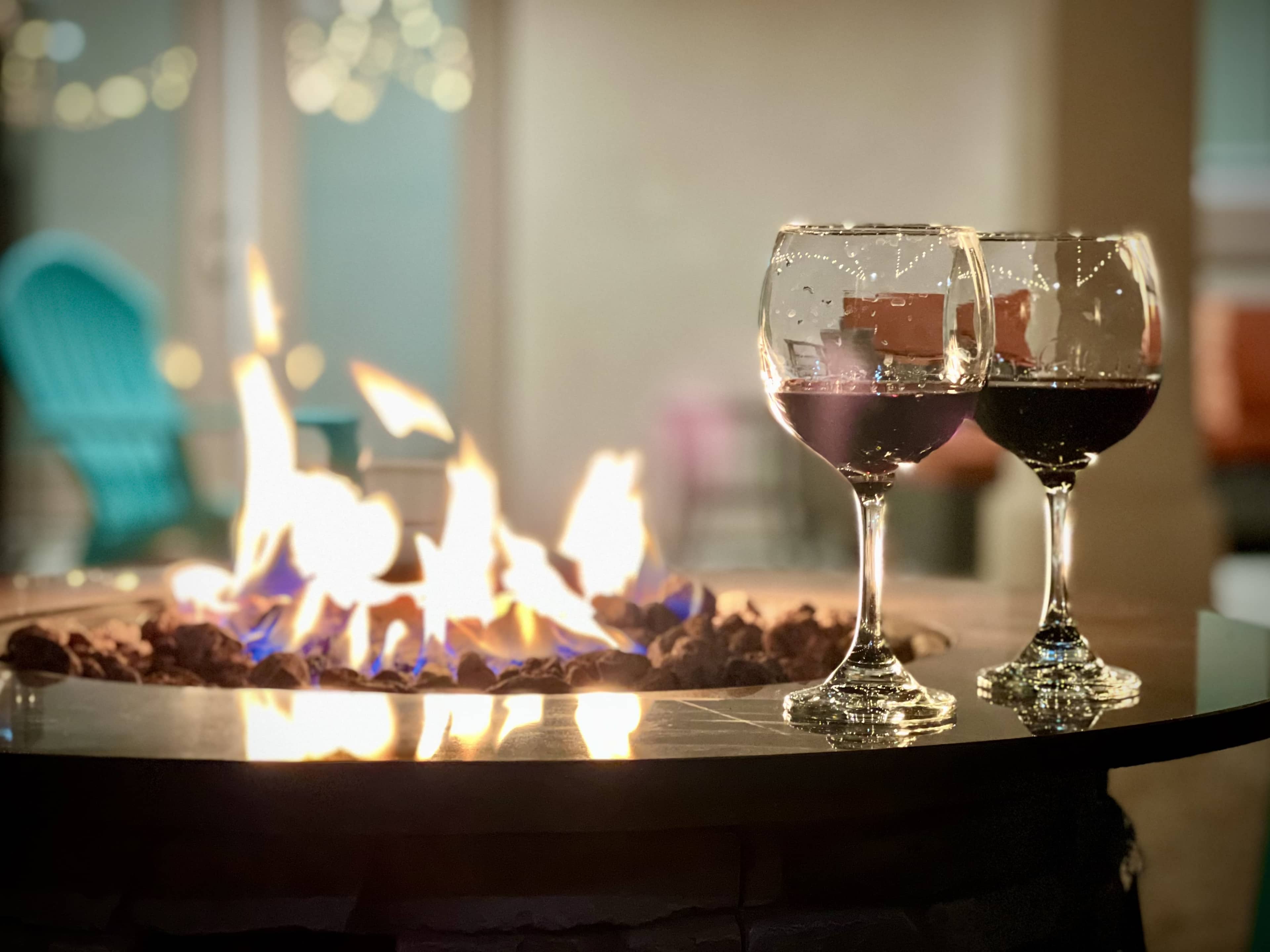 Two glasses of red wine are positioned on a table next to a glowing fire pit.