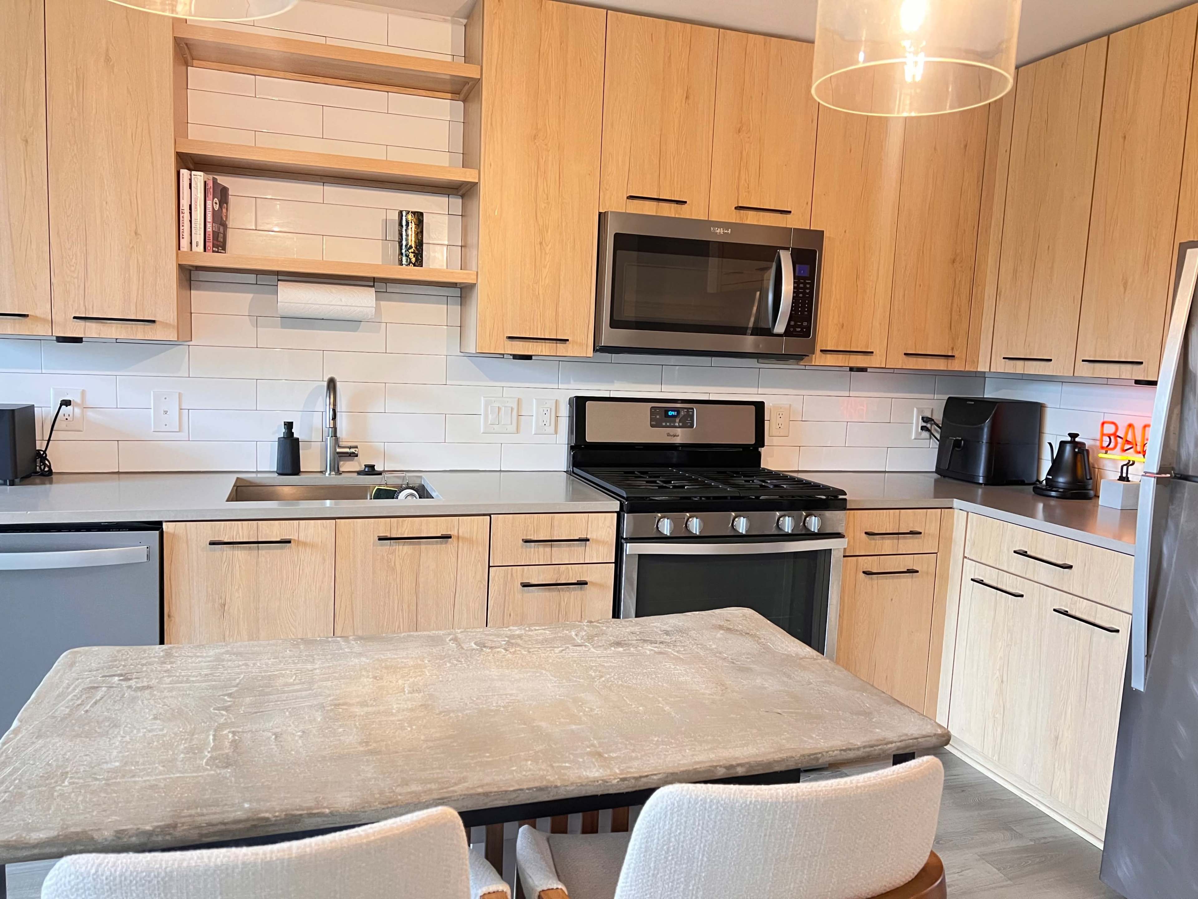 The image shows a modern kitchen featuring wooden cabinets, stainless steel appliances, and a small dining table with two chairs.