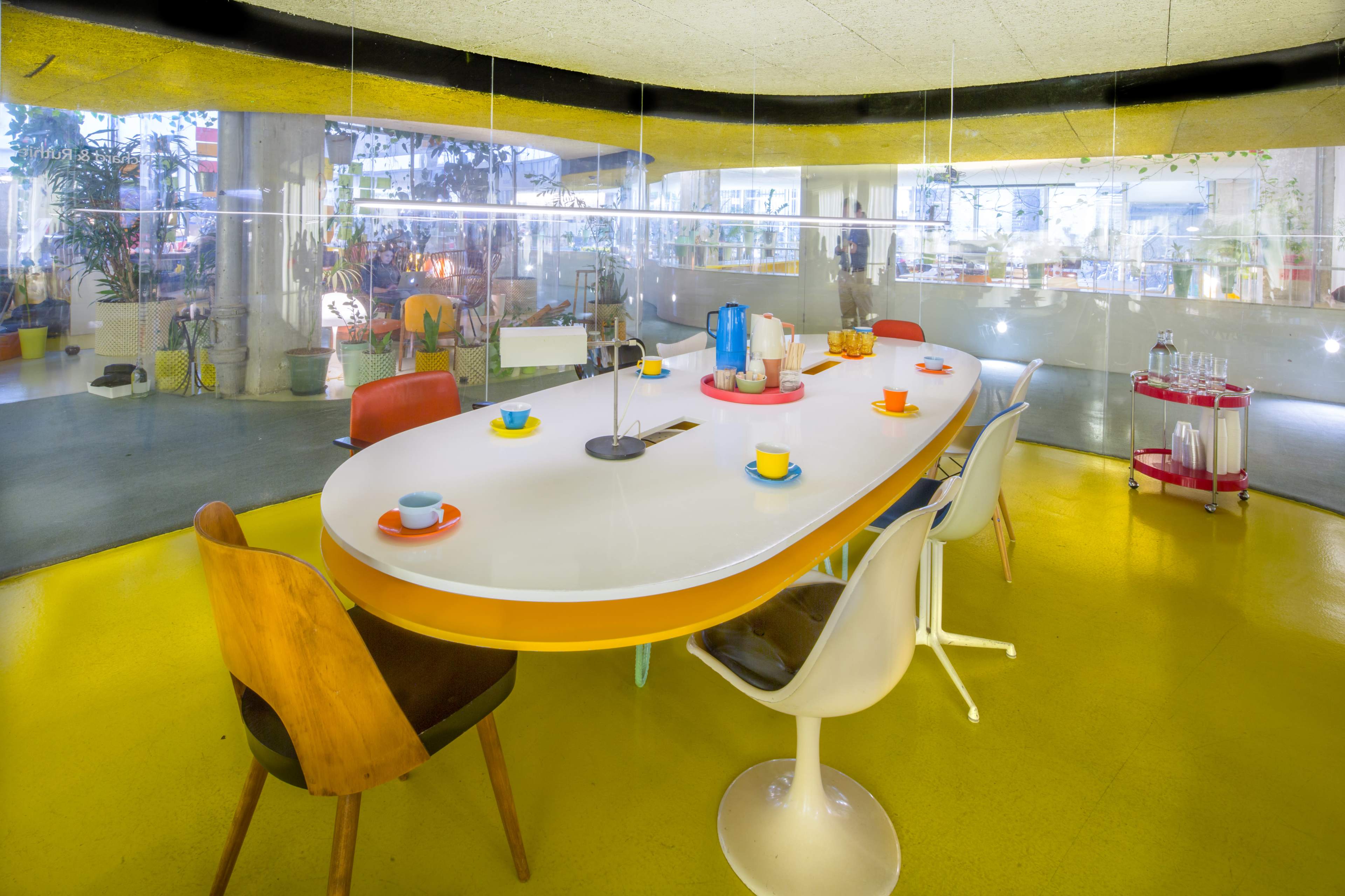 The image shows a modern dining area with a large, oval white table surrounded by colorful chairs on a bright yellow floor.