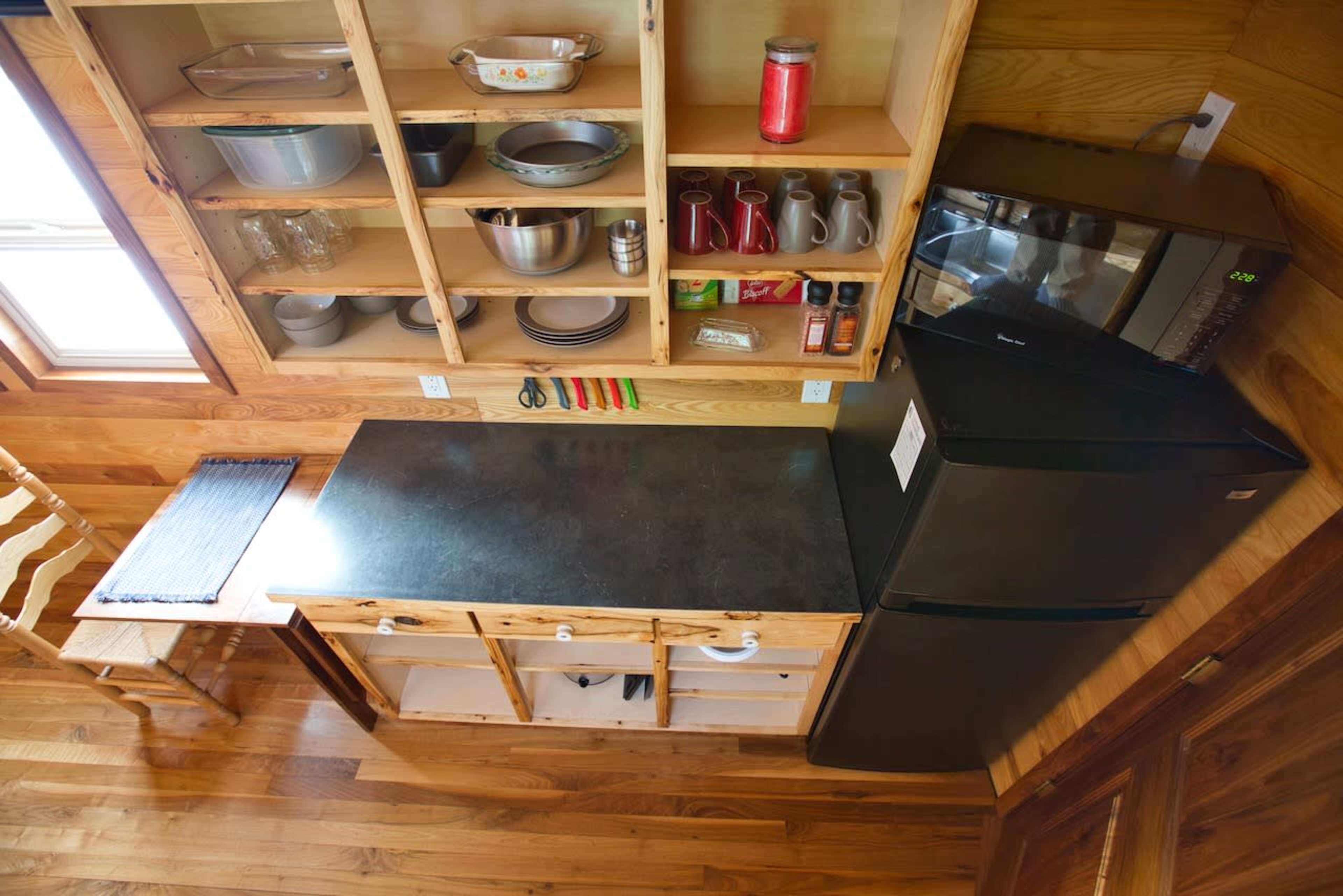 A compact kitchen with wooden shelves containing various kitchenware, a black countertop, a microwave, and a refrigerator.