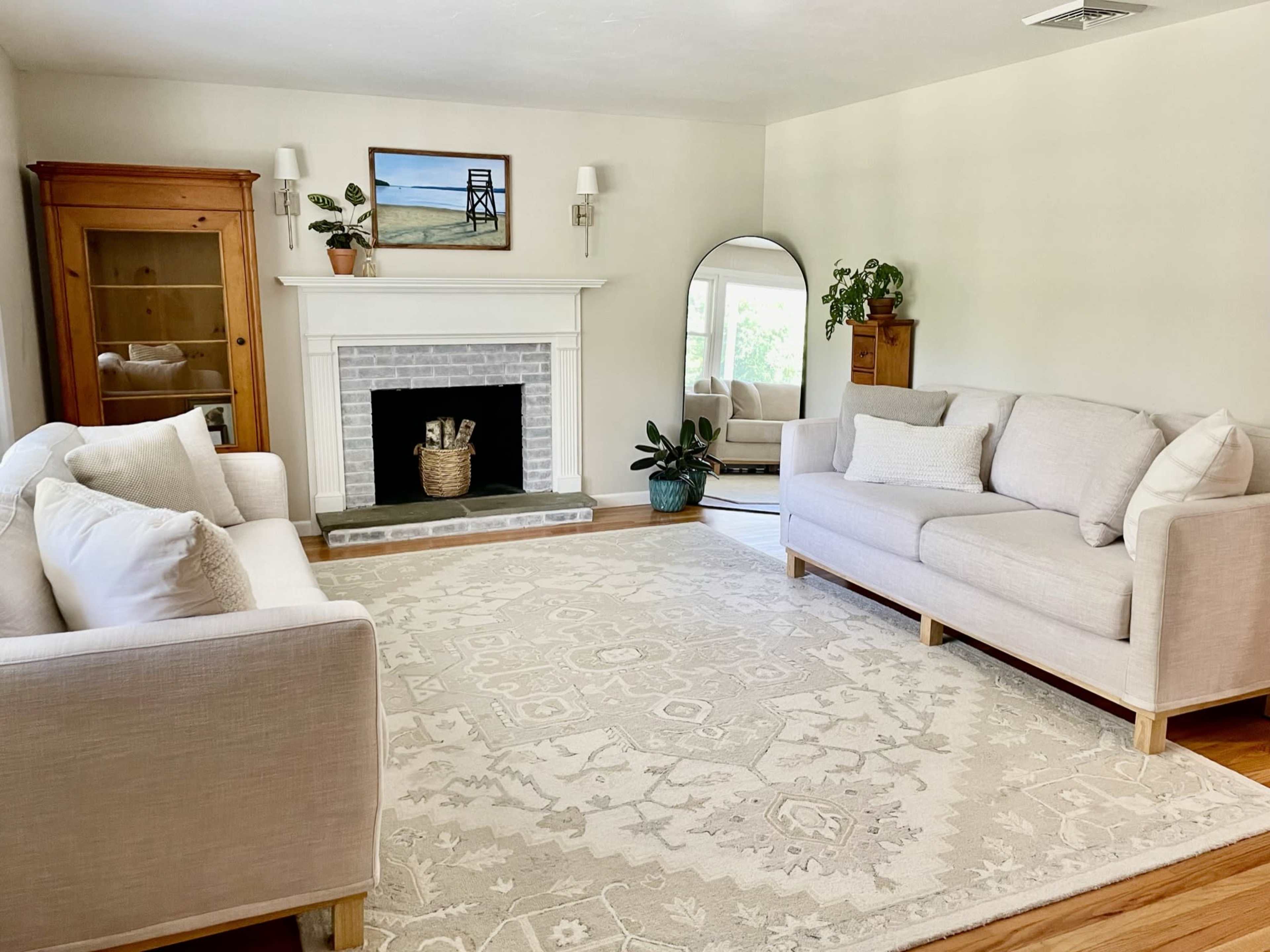 A light and airy living room features two beige sofas facing a fireplace, a large mirror on one wall, and a window allowing natural light to enter.