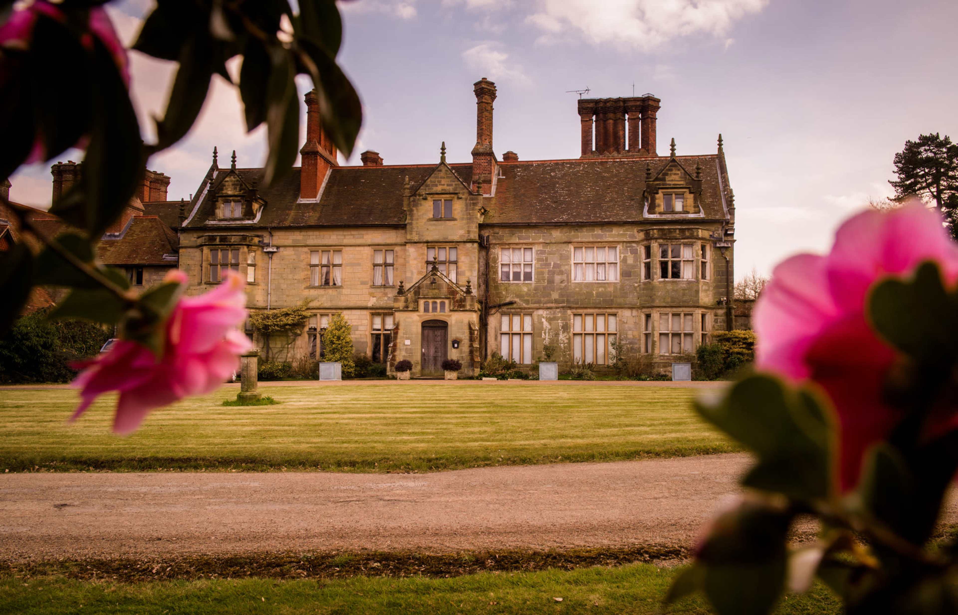 A large, historic mansion with intricate architecture is framed by blooming flowers in the foreground and sits on a manicured lawn.