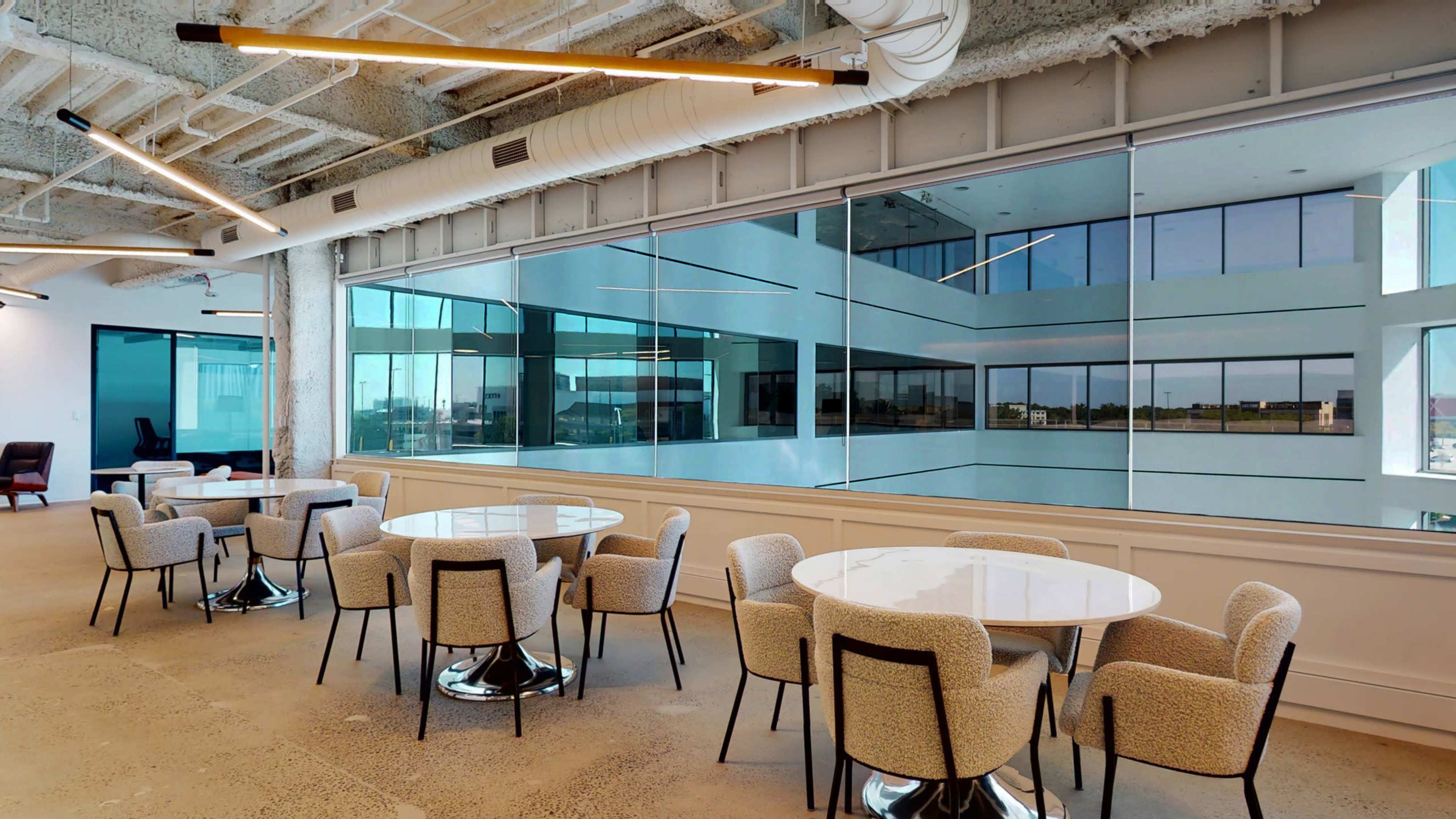 A modern office lounge area features several round tables surrounded by upholstered chairs, offering a view through large windows overlooking an indoor atrium.