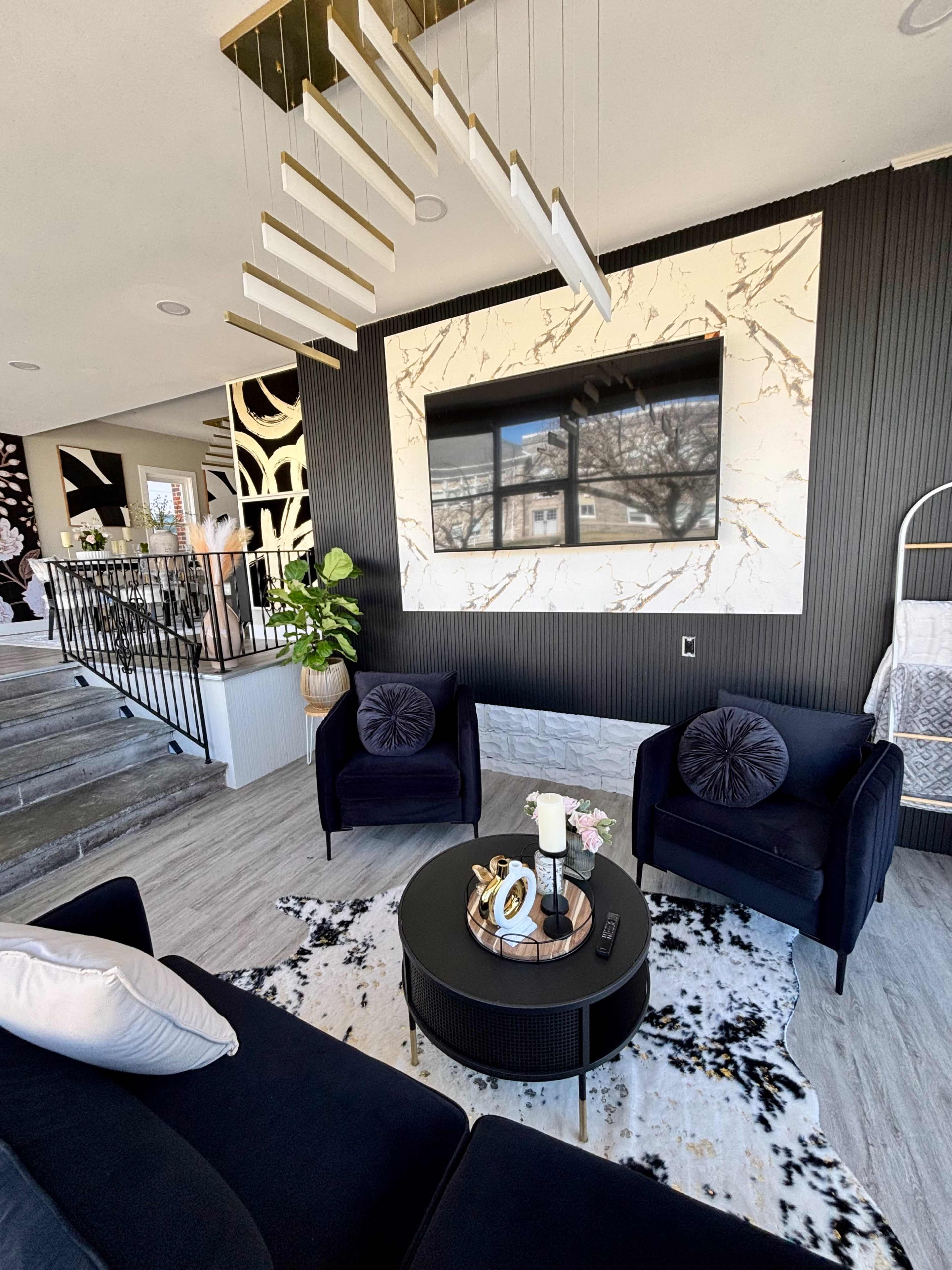 A modern living room features two velvet chairs facing a round coffee table, while a large TV is mounted on a marble-patterned wall behind them.