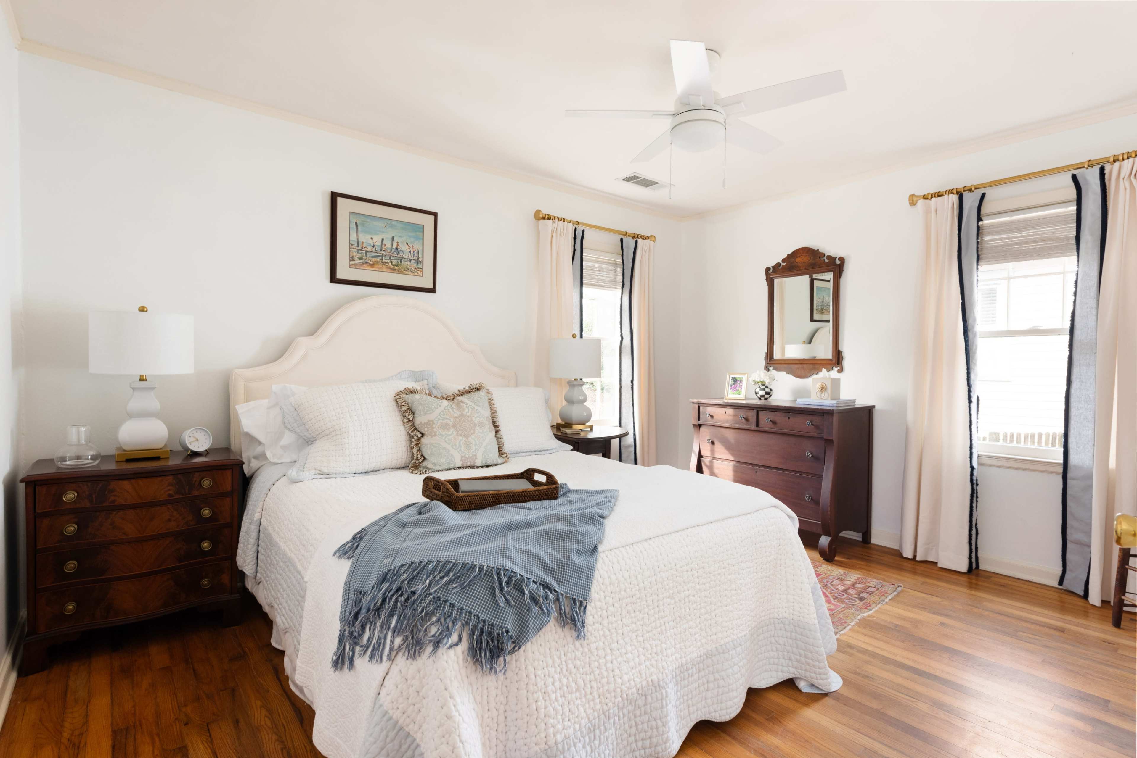 The image shows a well-furnished bedroom featuring a bed with a light-colored quilt, a wooden dresser, and natural light coming through a window.