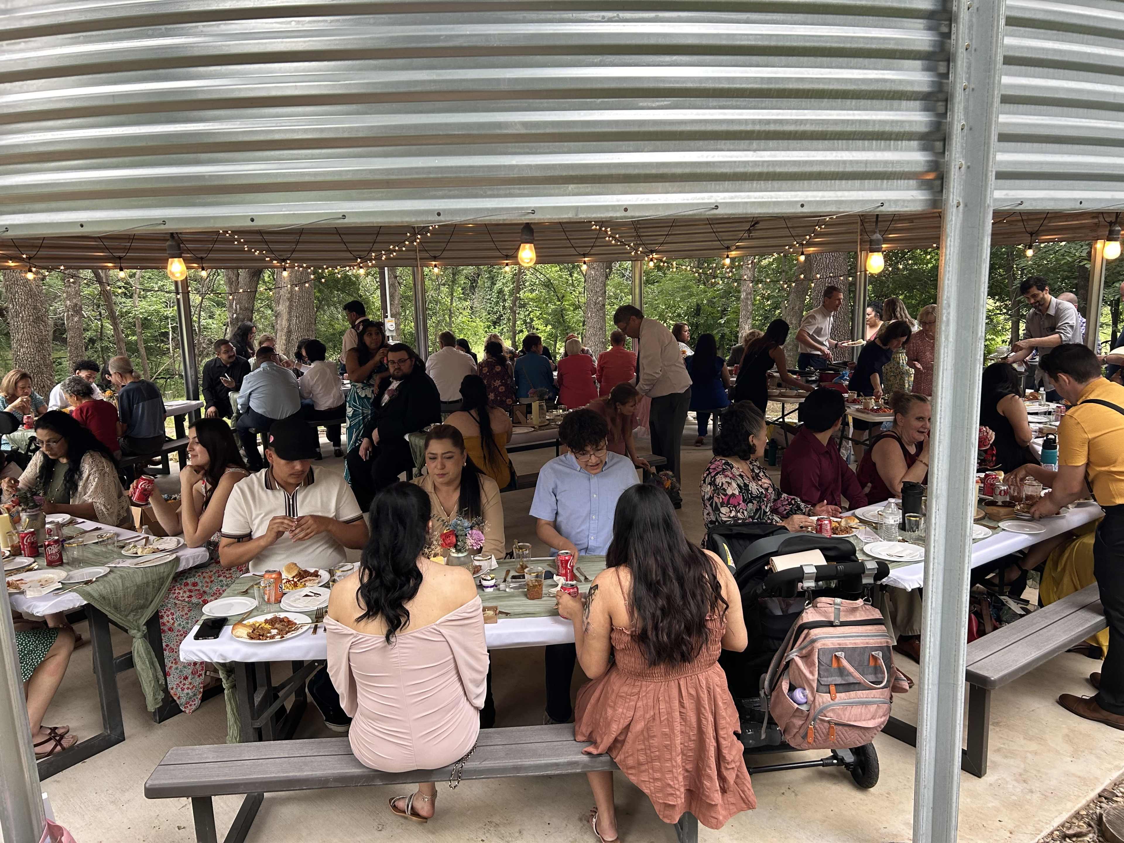 A large group of people gathers around several tables in a covered outdoor space, enjoying a meal amidst trees and string lights.