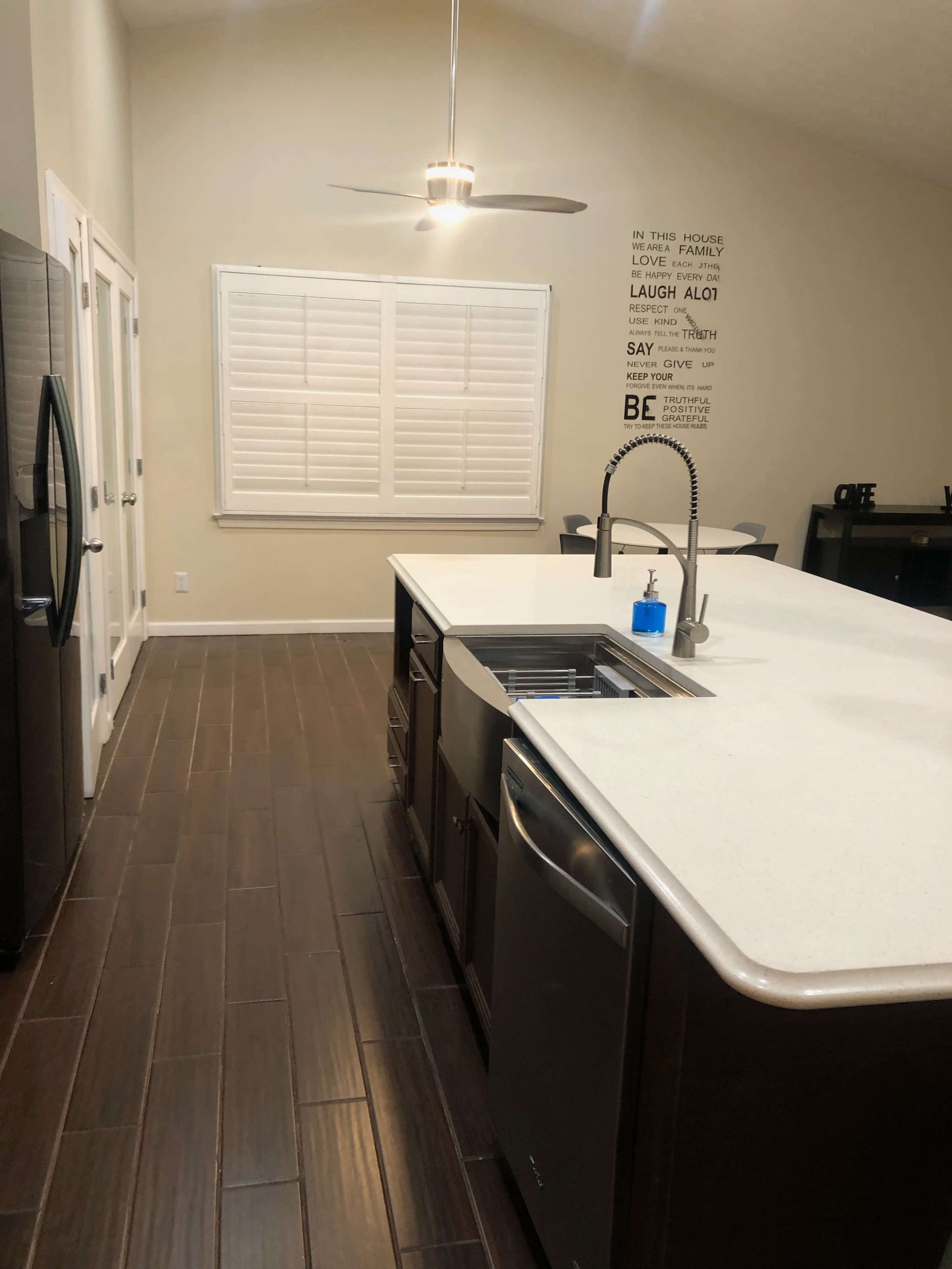 A modern kitchen with dark wood flooring, a central island with a sink, and a large window equipped with white shutters.
