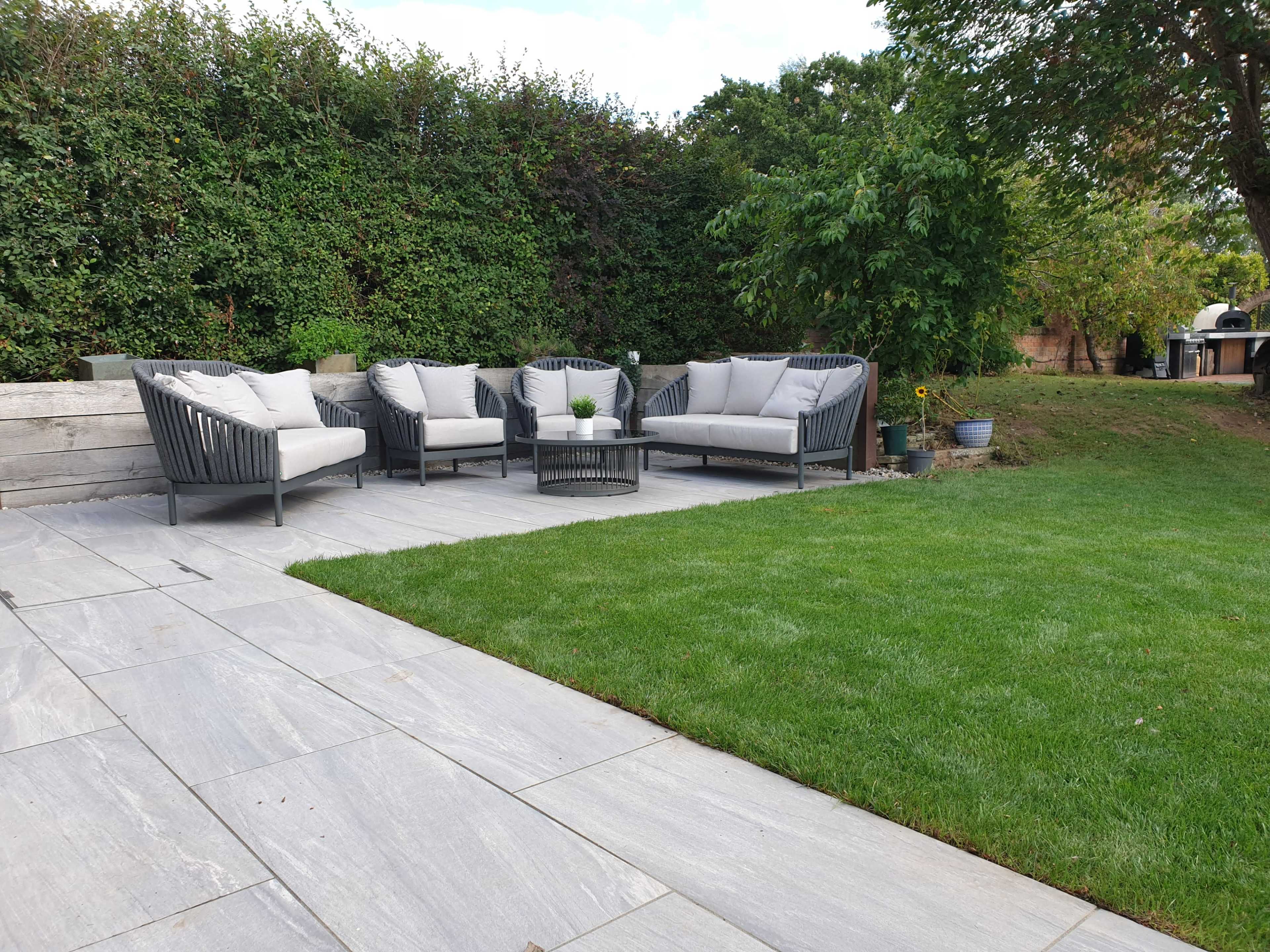 A patio area with gray stone flooring, a set of four wicker chairs with cushions, and a small table, surrounded by green grass and shrubs.
