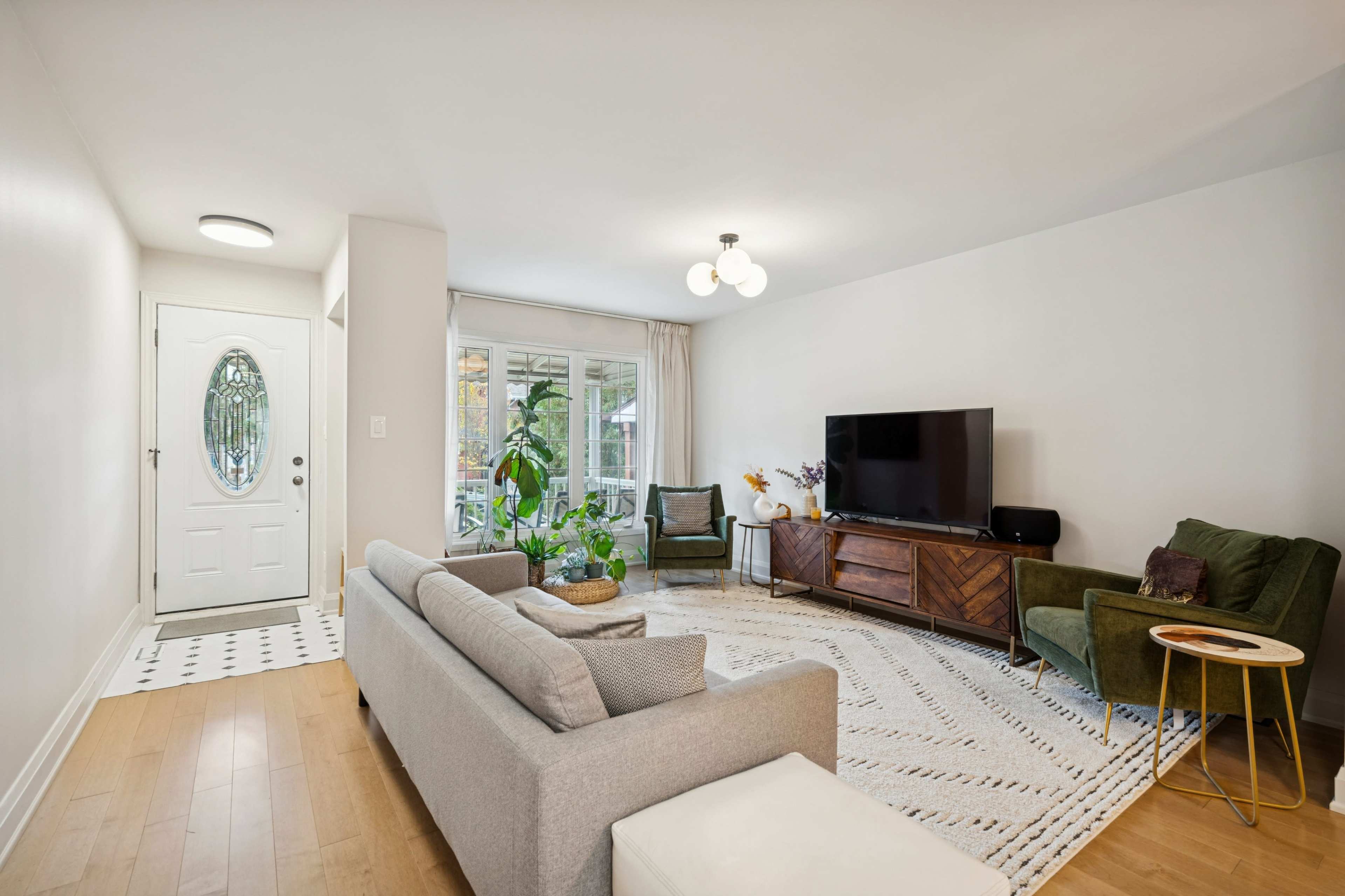 The living room features a modern design with a gray sofa, a large television on a wooden stand, two armchairs, and a round rug.