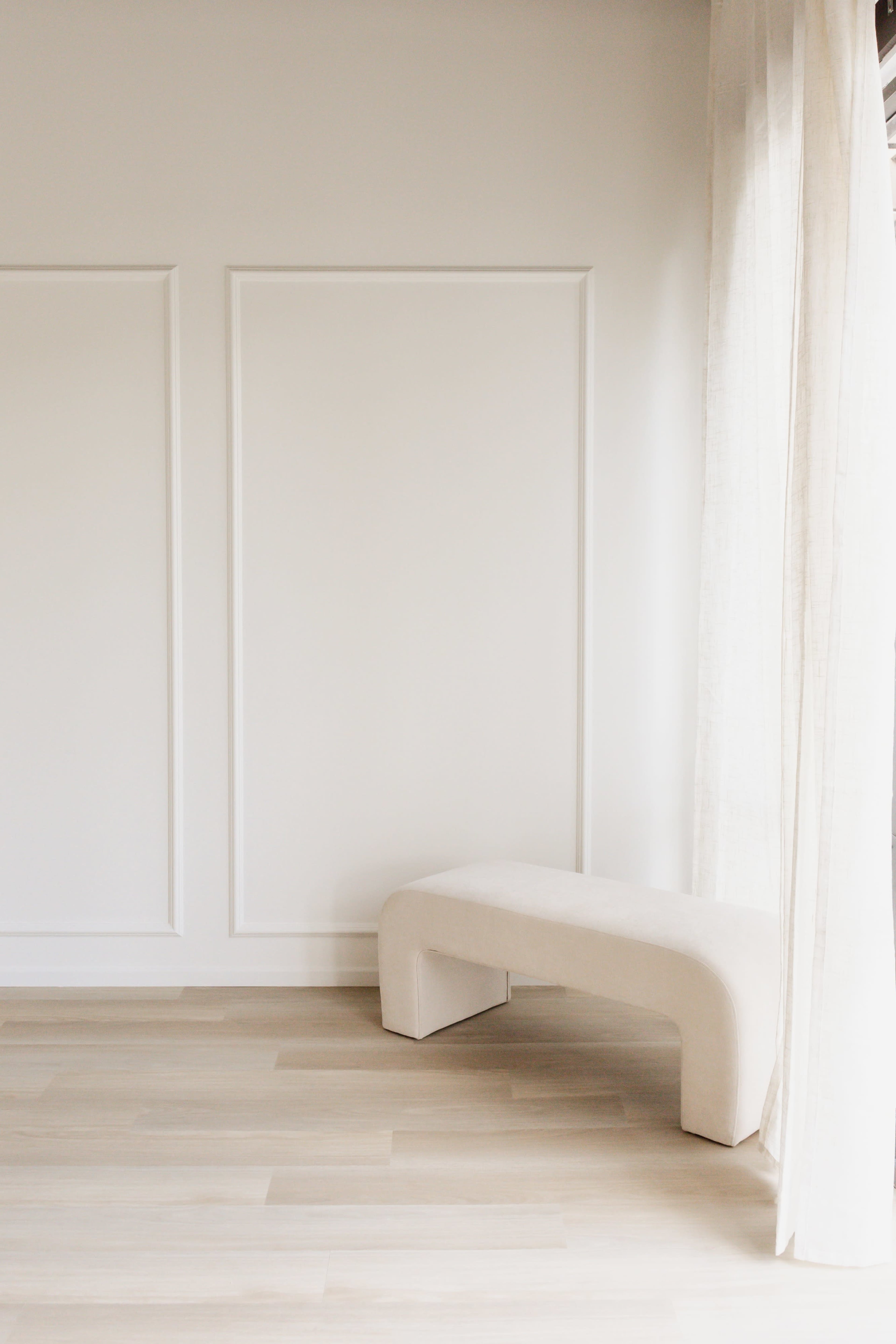 The image shows a minimalist interior with a curved, light-colored bench positioned against a plain wall, accompanied by sheer curtains.
