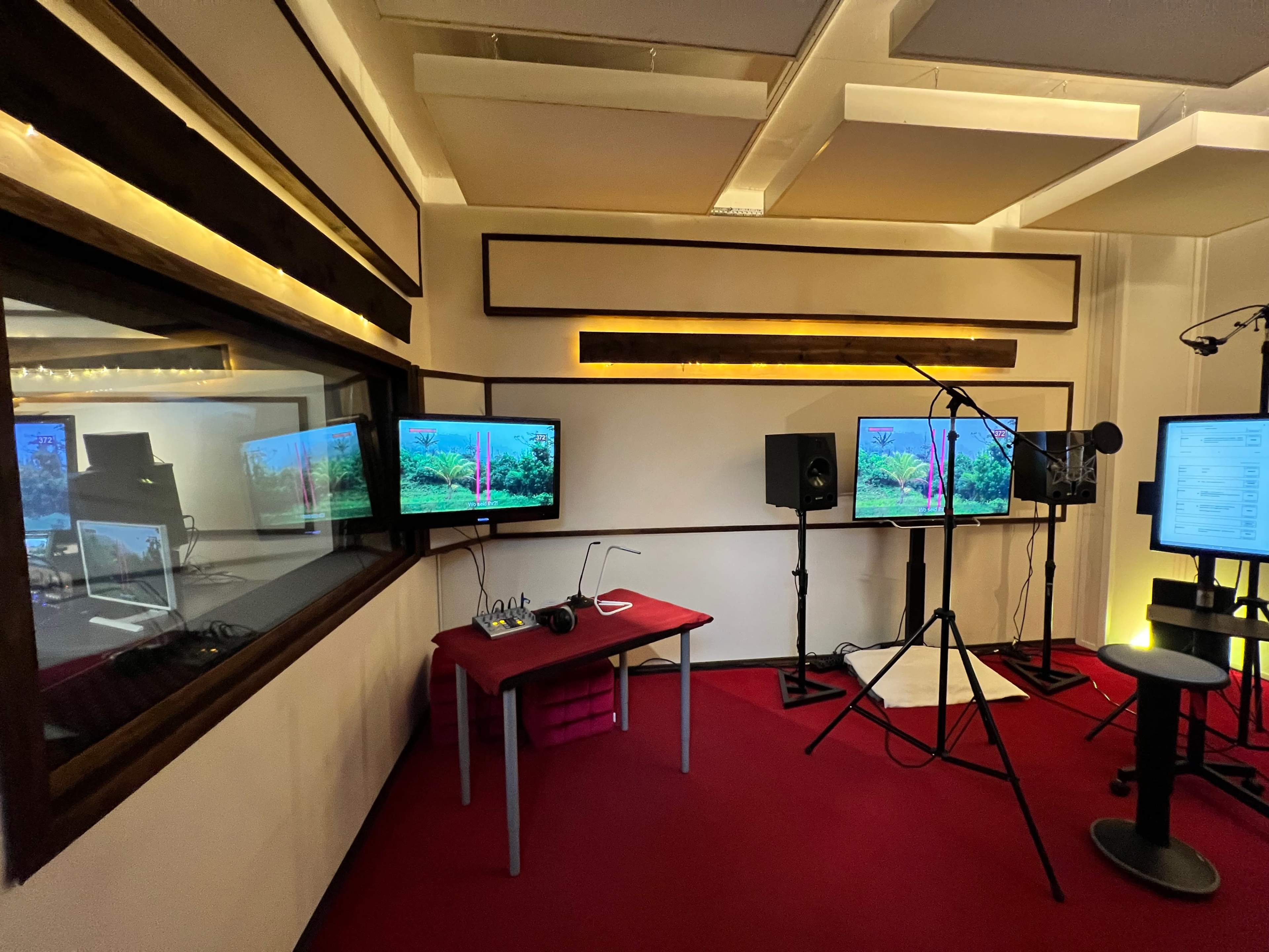 A recording studio with two monitors displaying a nature scene, surrounded by audio equipment and a red carpeted floor.